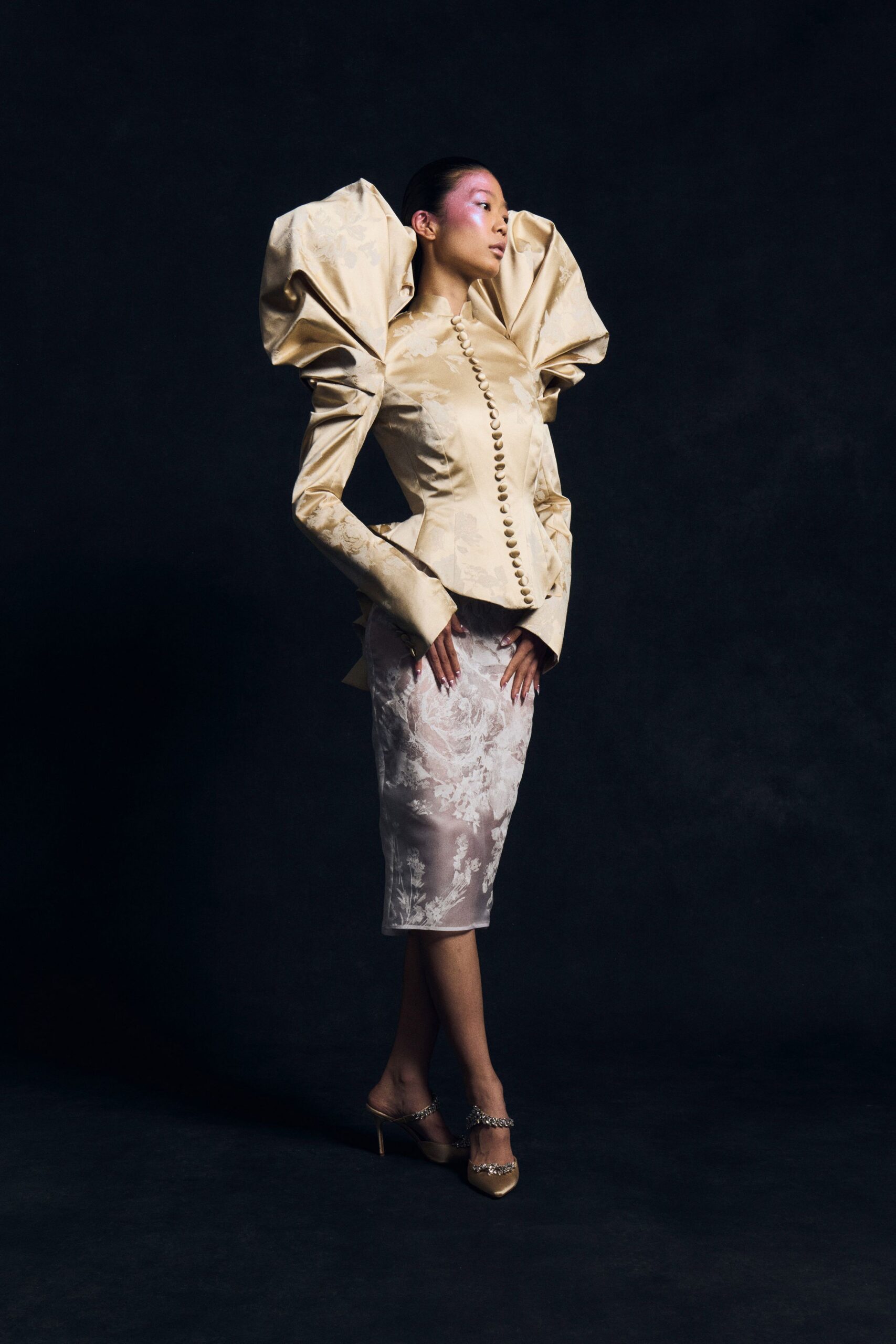 Champagne jacquard jacket, giant sculpted sleeves and a row of fabric buttons. Pale fitted skirt in sheer floral brocade, ending just below the knee. Pointed satin mules with crystal chain straps. For cutting the cake in a ballroom where the flowers are already wilting.