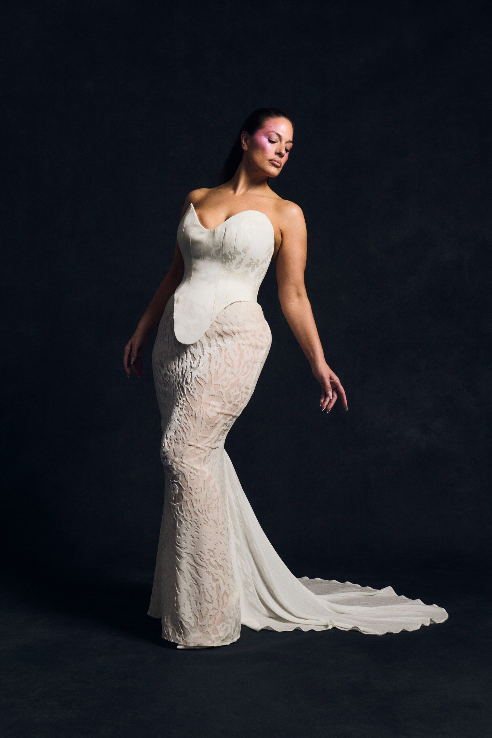 Ivory strapless mermaid gown with a heart-cut corset and floral jacquard. Sheer back train spilling from the hips, hem pooling in a long oval. After the chapel empties, standing alone on cold theater carpet.