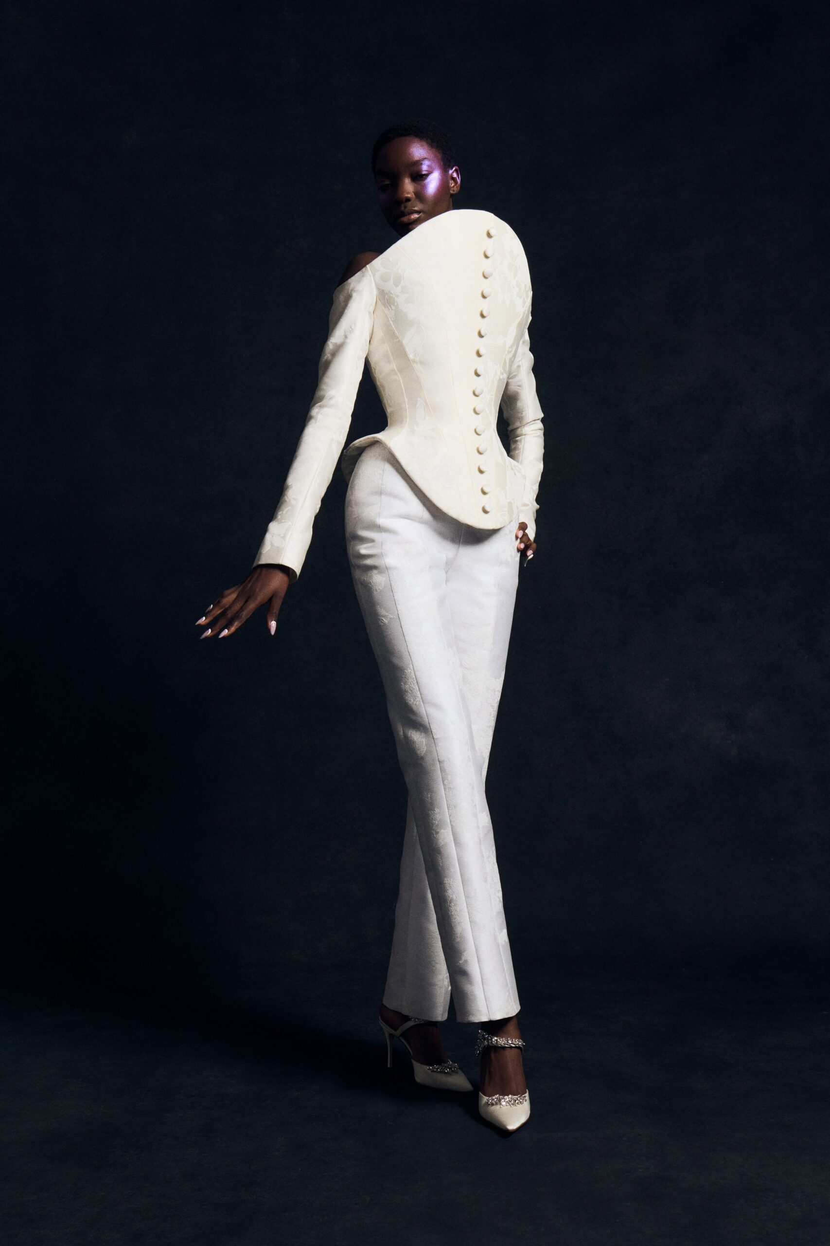 Ivory jacquard corset top, one shoulder bare, covered buttons down the back. Matching bootcut trousers in the same floral jacquard, pressed through the knee. Ivory slingback pumps with crystal trim at toe and strap. After the ceremony, crossing a hotel ballroom before the glasses are cleared.