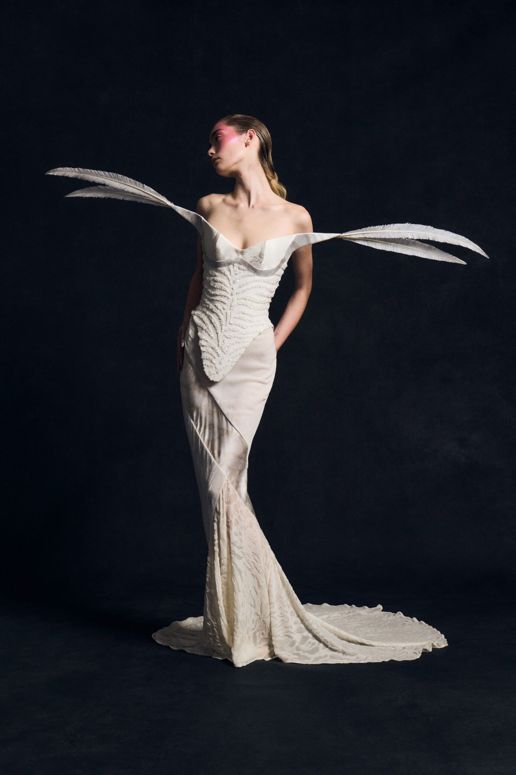 Ivory mermaid gown with a bead-ridged corset and pointed basque front. Two long white feather plumes springing straight from the off-shoulder neckline. Jacquard train pooling out behind in a faint animal-like pattern. After midnight at the natural history museum, alone with the bird cases.