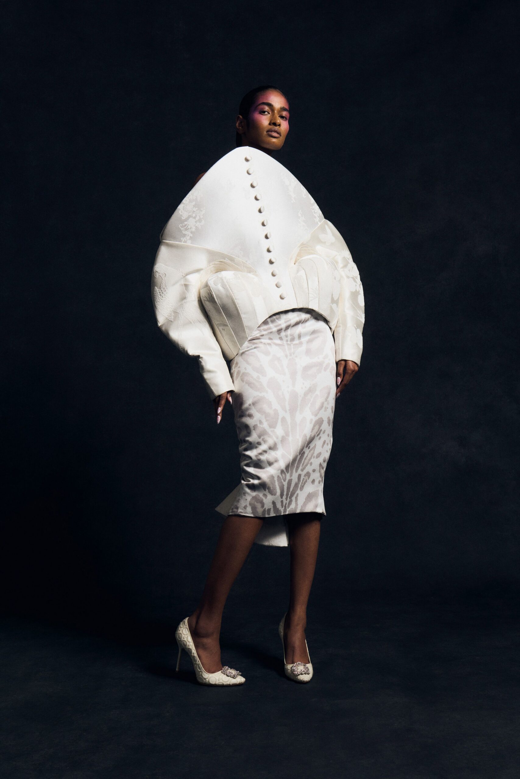 Ivory sculpted jacket ballooning at the sleeves, domed buttons climbing the front. Pearled satin pencil skirt with a pale organic motif and slight fishtail kick. Ivory brocade pumps topped with crystal buckle clusters. City hall wedding, then champagne in a room with bad carpet.