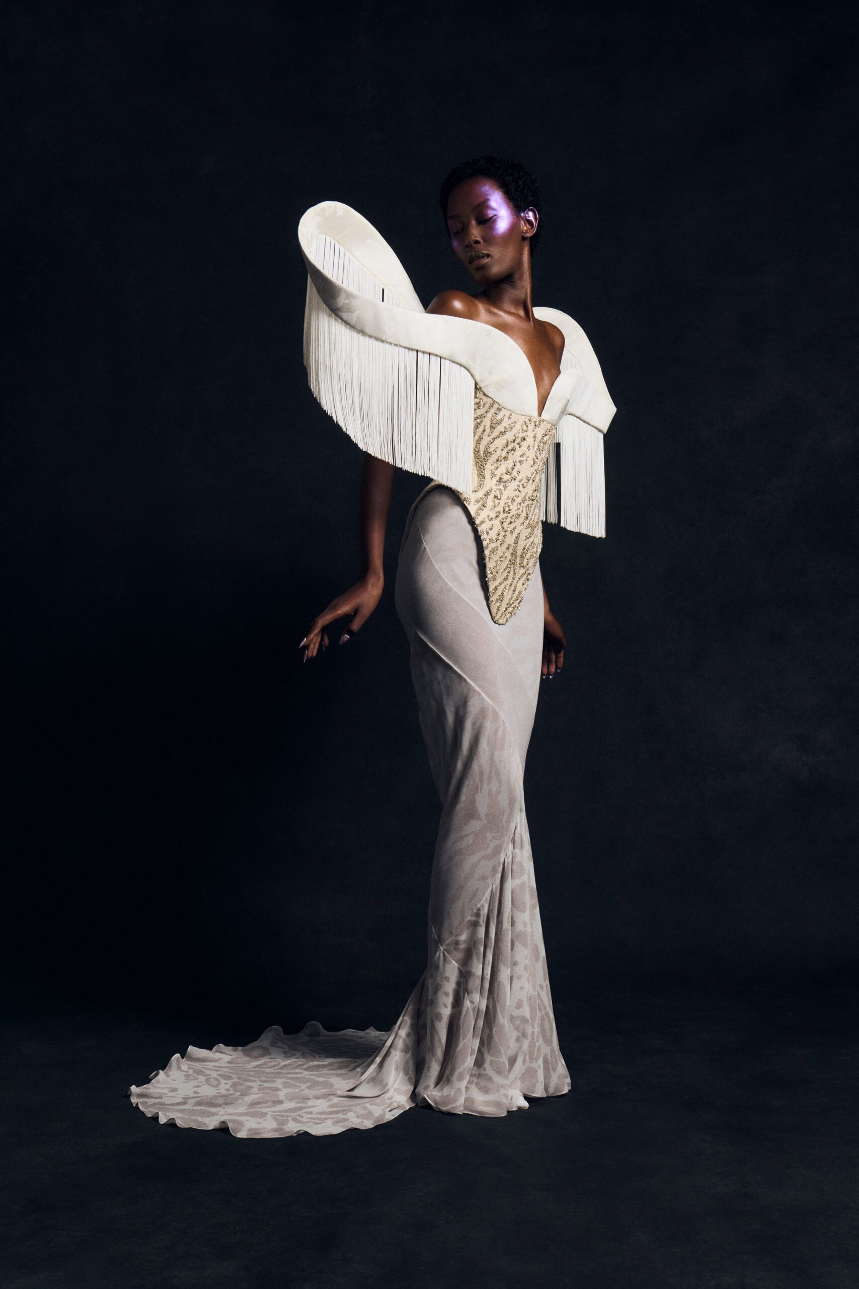 Ivory sculpted satin scrolls, hung with long curtain fringe. Gold beaded corset tapering to a sharp front point. Pearl grey column in sheer devore, train puddling into mottled spots. Opera-house stairwell after midnight, the marble still cold through the train.