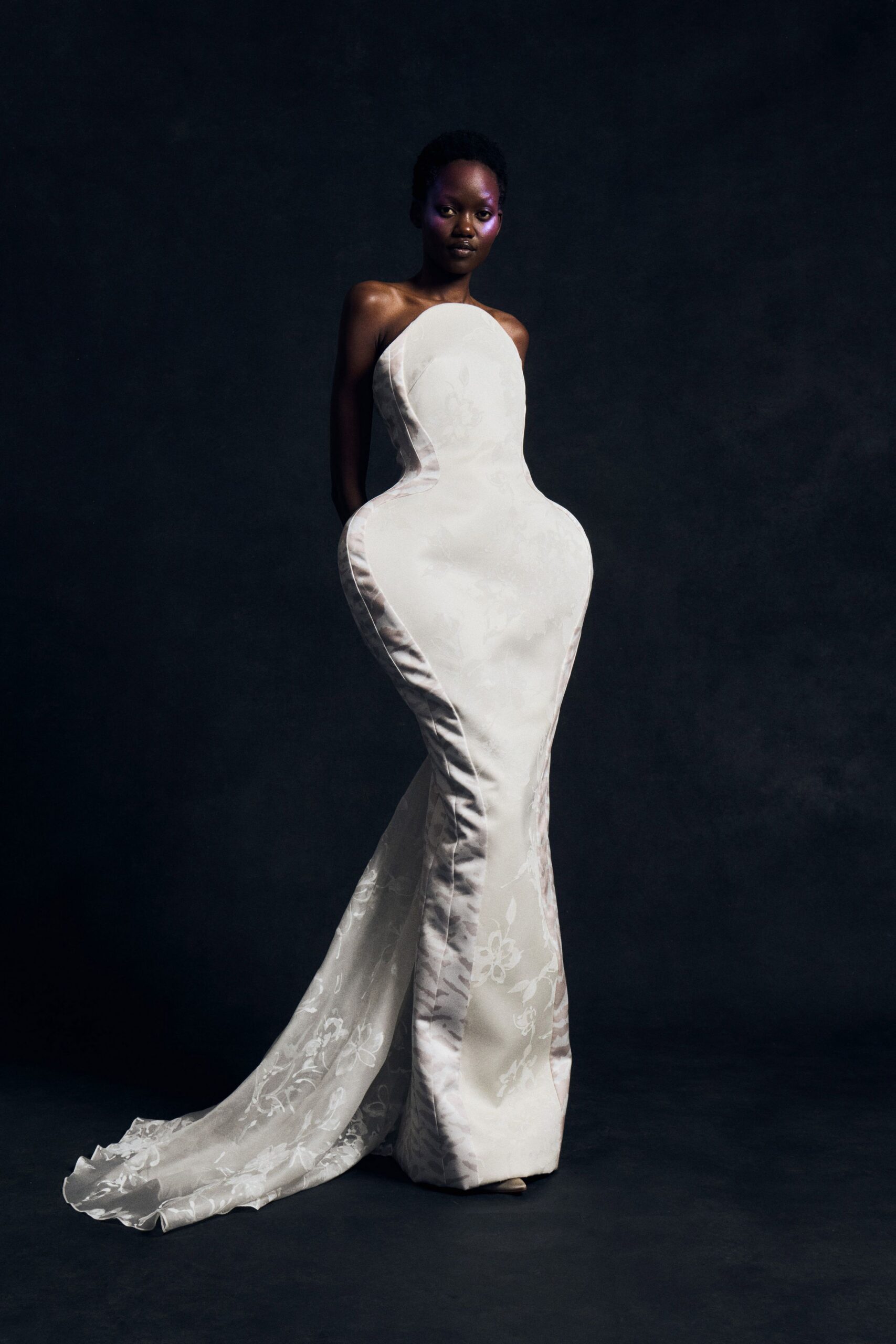 Strapless ivory satin gown, floral jacquard panels swelling into exaggerated hips and a trailing fishtail. Pale pointed shoes barely peeking beneath the hem. After the vows, crossing a cold ballroom before the lights come up.