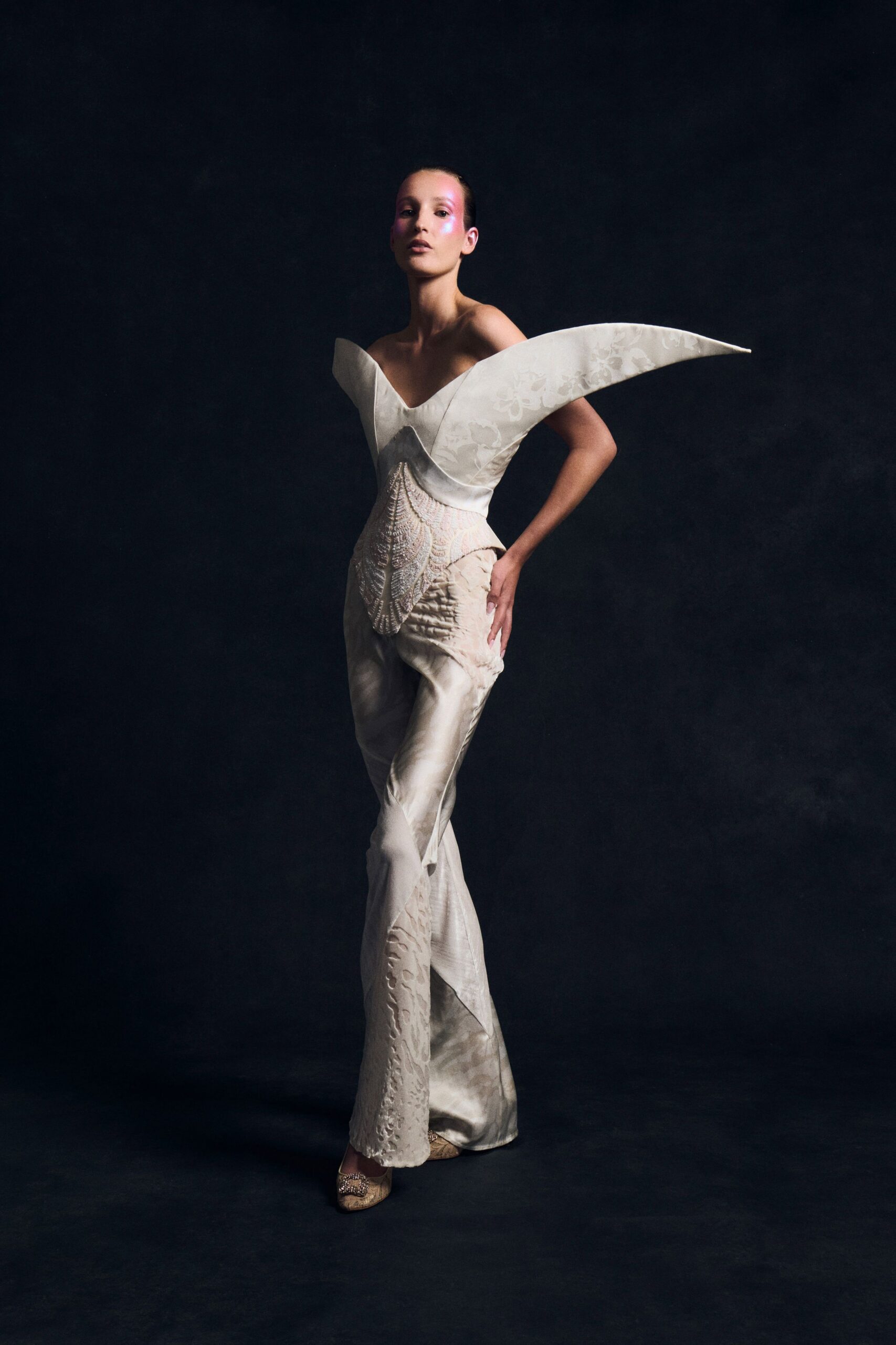 Ivory satin bodice with winged off-shoulder points and faint floral jacquard. Shell-pink beaded panel cupping the waist like a stitched carapace. Pearl flared trousers in liquid satin, laser-cut motifs climbing one leg. Brooched pumps in pale metallic brocade. For arriving after midnight at a crumbling seaside ballroom.