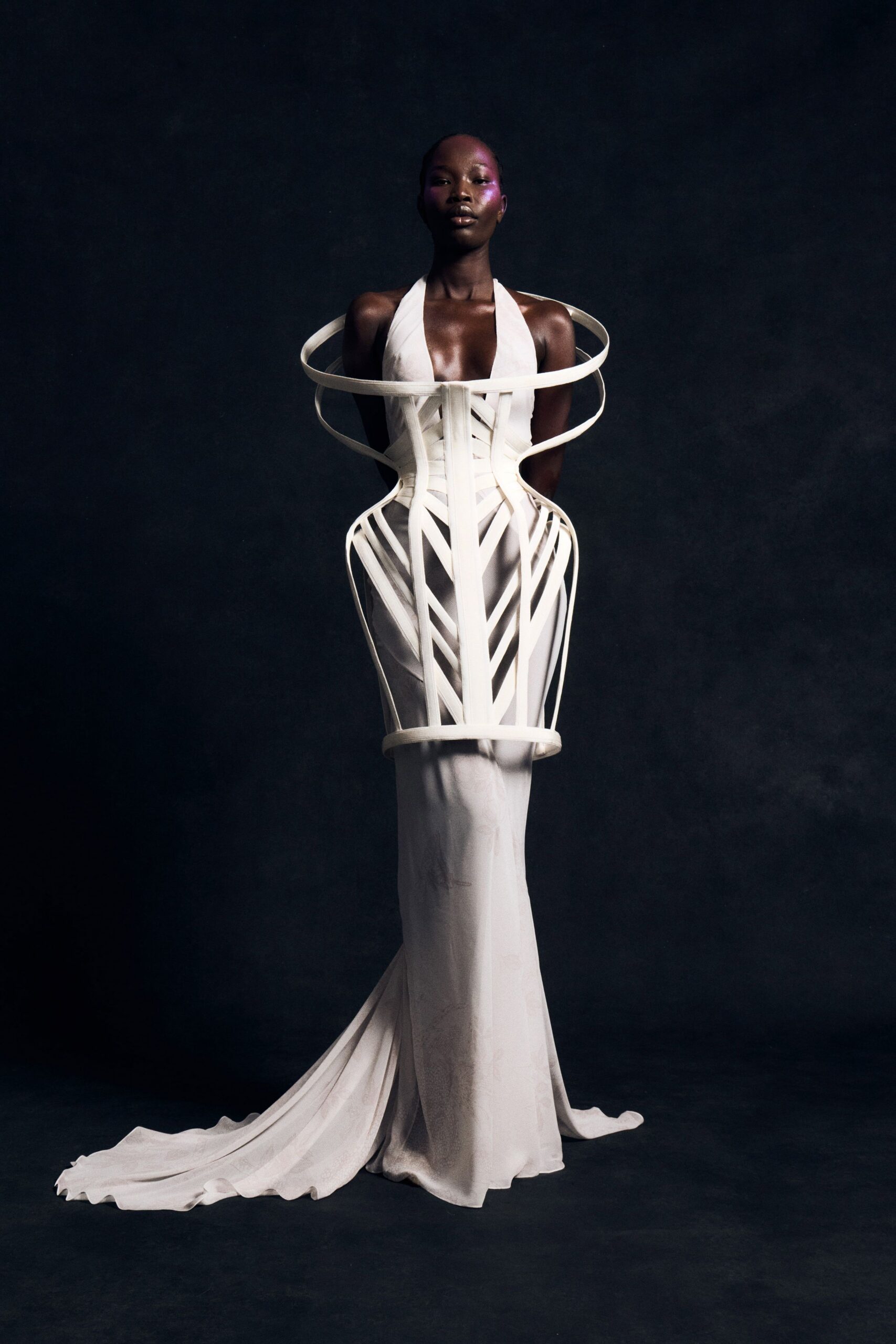 Ivory halterneck column gown in liquid satin, pooling into a long train. White ribbon cage corset circling the shoulders, chevrons framing the hips. For standing in the museum after closing, still roped off from touch.