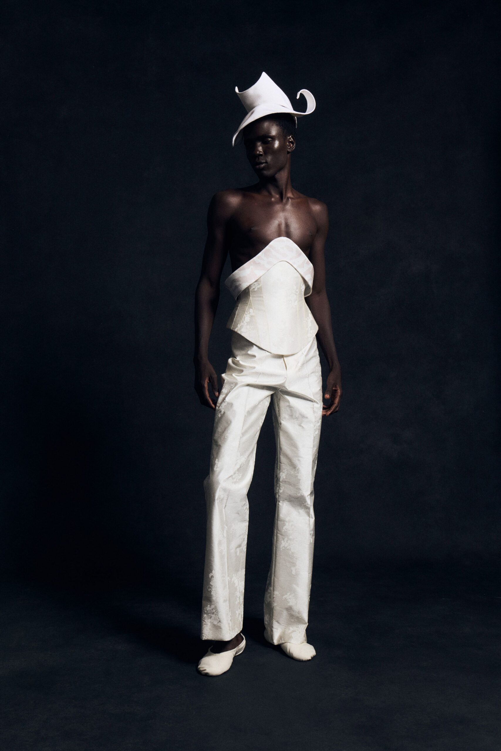 Ivory felt hat, brim curling up into two horned points. Strapless ivory brocade bustier with a folded band crossing the bust. Straight ivory brocade trousers, pressed flat through the front. Soft ivory slipper flats. Civil ceremony at noon, the courthouse steps still wet from rain.