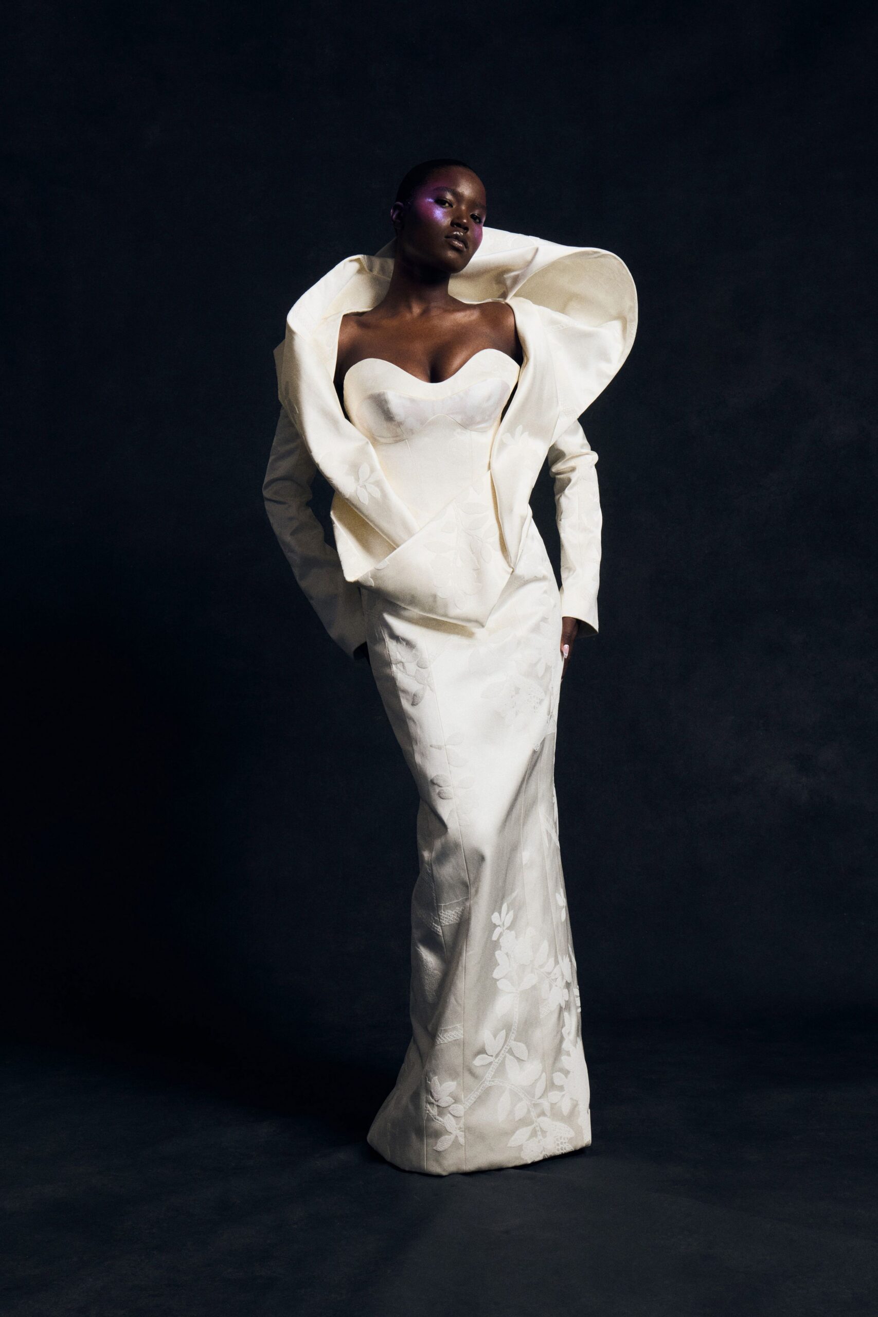 Ivory satin column gown with a sweetheart bustier and scattered floral appliqué. Matching cropped jacket thrown open, its collar rolled into a huge sculptural loop. Town hall wedding, the florist buckets still sweating by the side door.