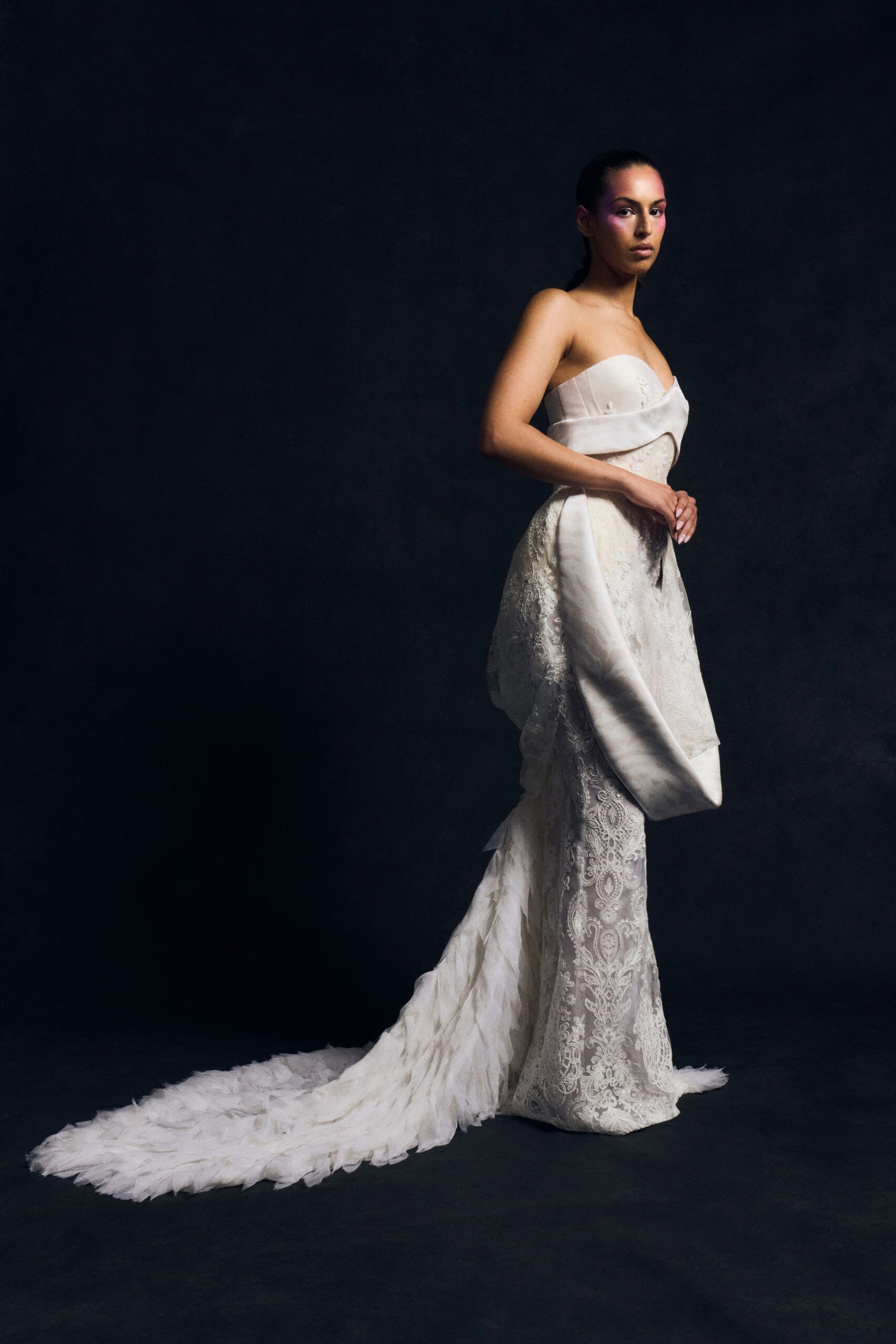 Strapless ivory gown with a folded satin band crossing the bust. Sheer lace column skirt under a draped overskirt panel. Long chiffon train, softly crumpled into feathered tiers at the hem. After the chapel, standing still while the grass stains the underside.