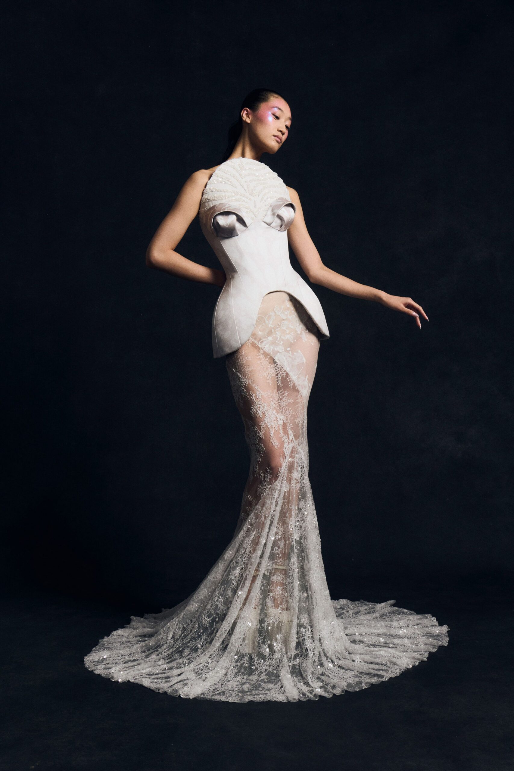 Ivory satin corseted bodice with sculpted cups and a split peplum. Sheer fishtail lace skirt, sequins catching through the pooled train. Beaded high-neck panel ruched into shell-like arcs above the bust. For the first fitting after the cathedral bells have started.