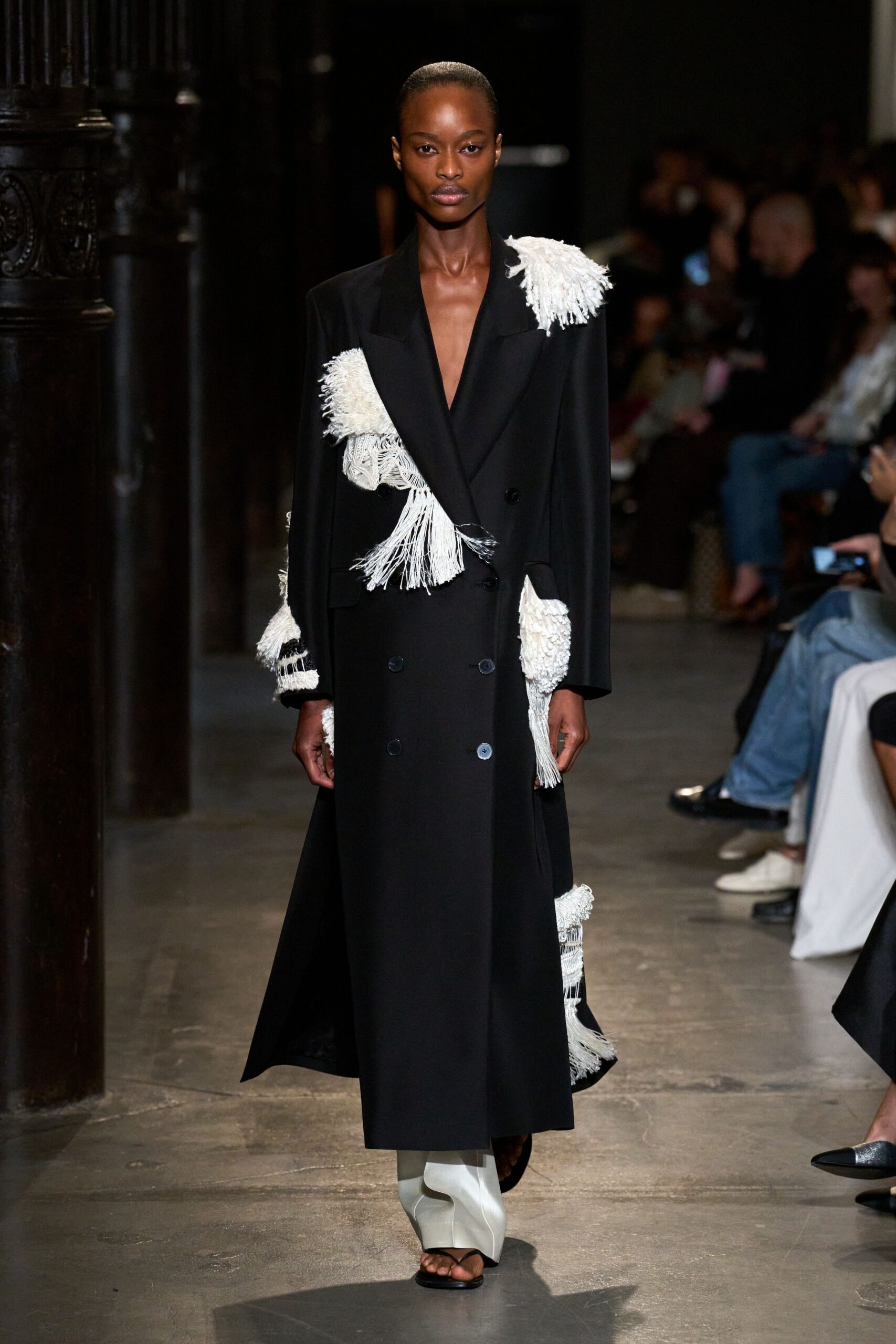 Black double-breasted coat with oversized peaked lapels and raw ivory fringe appliques. White leather boots split open at the toe over black thong sandals. Crossing a marble lobby after midnight with confetti caught on your sleeve.