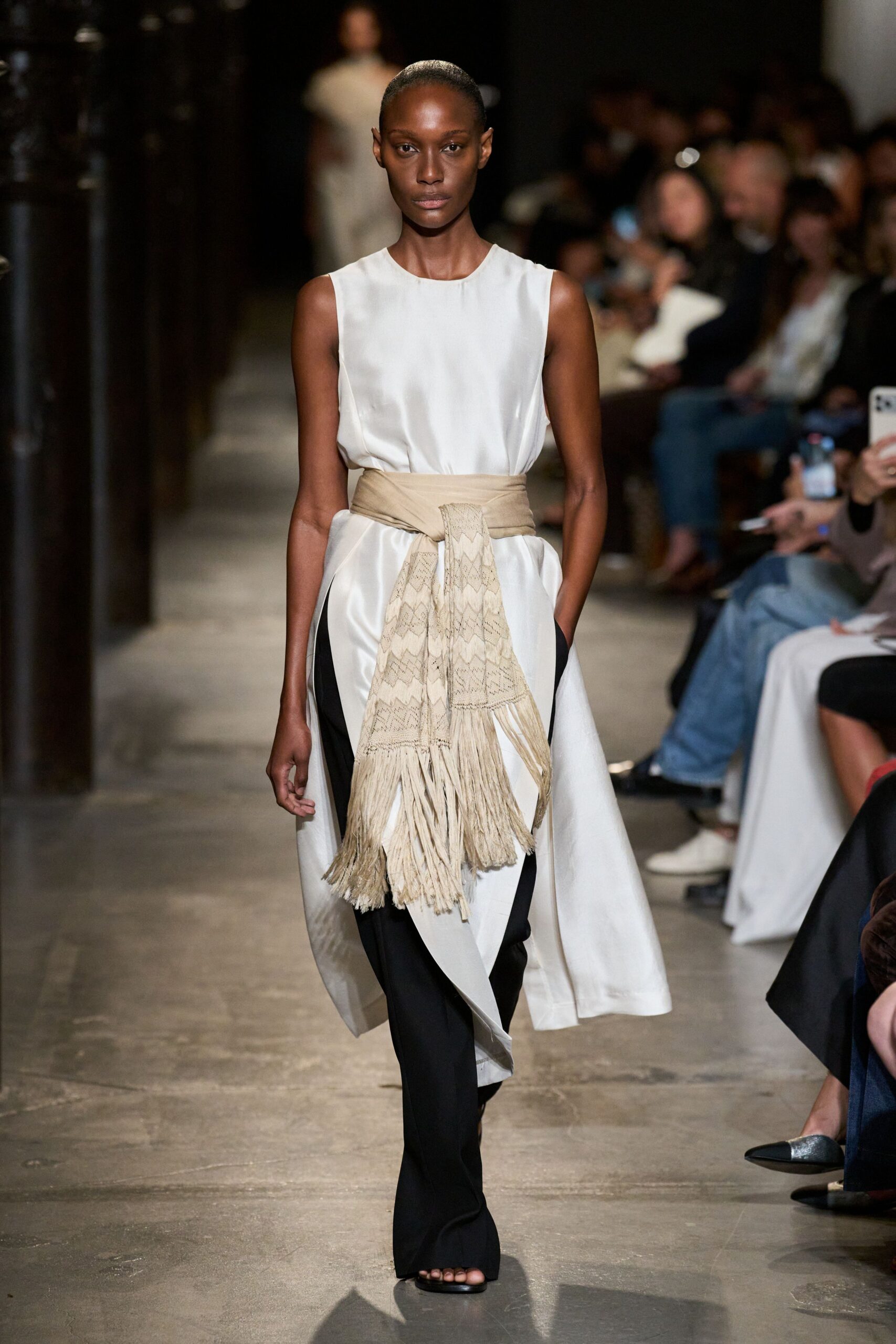 Ivory satin sleeveless dress, cut away at the sides into long panels. Wide sand wrap belt with a loosely knotted fringe-ended woven scarf. Black straight trousers, visible through the dress's split front. Black flat sandals, barely there under the hem. Crossing a cloister courtyard after dinner, scarf ends brushing the stone.