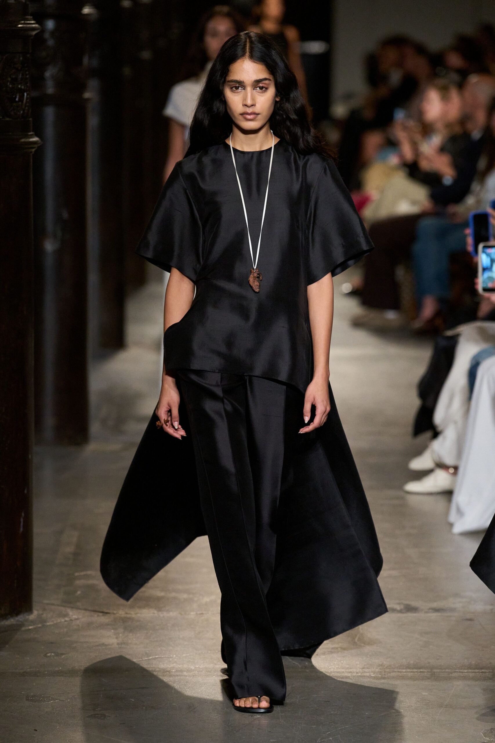 Long cord necklace with a small carved heart pendant. Black satin tunic with broad sleeves and a cutaway side. Floor-length back panel flaring from beneath the tunic. Wide black trousers with a pressed front crease. Barely-there black thong sandals. Crossing a stone cloister just after the bells.