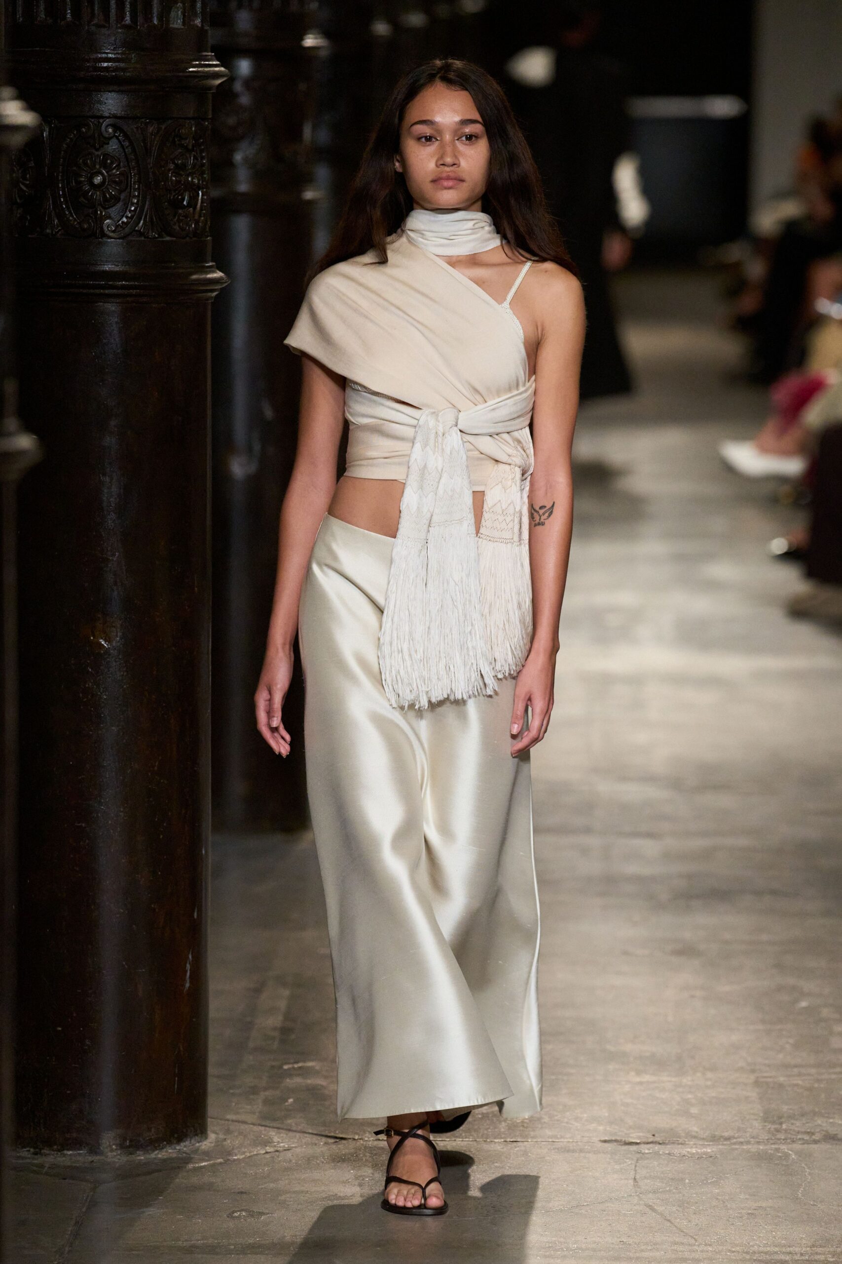 Cream jersey wrap top, one-shoulder and knotted with a long fringed panel. Matching jersey scarf looped close at the neck. Pearl satin column skirt cut low on the hip with liquid shine. Black thong sandals, barely there straps. Crossing a hotel corridor before dawn, silk hem skimming the stone.