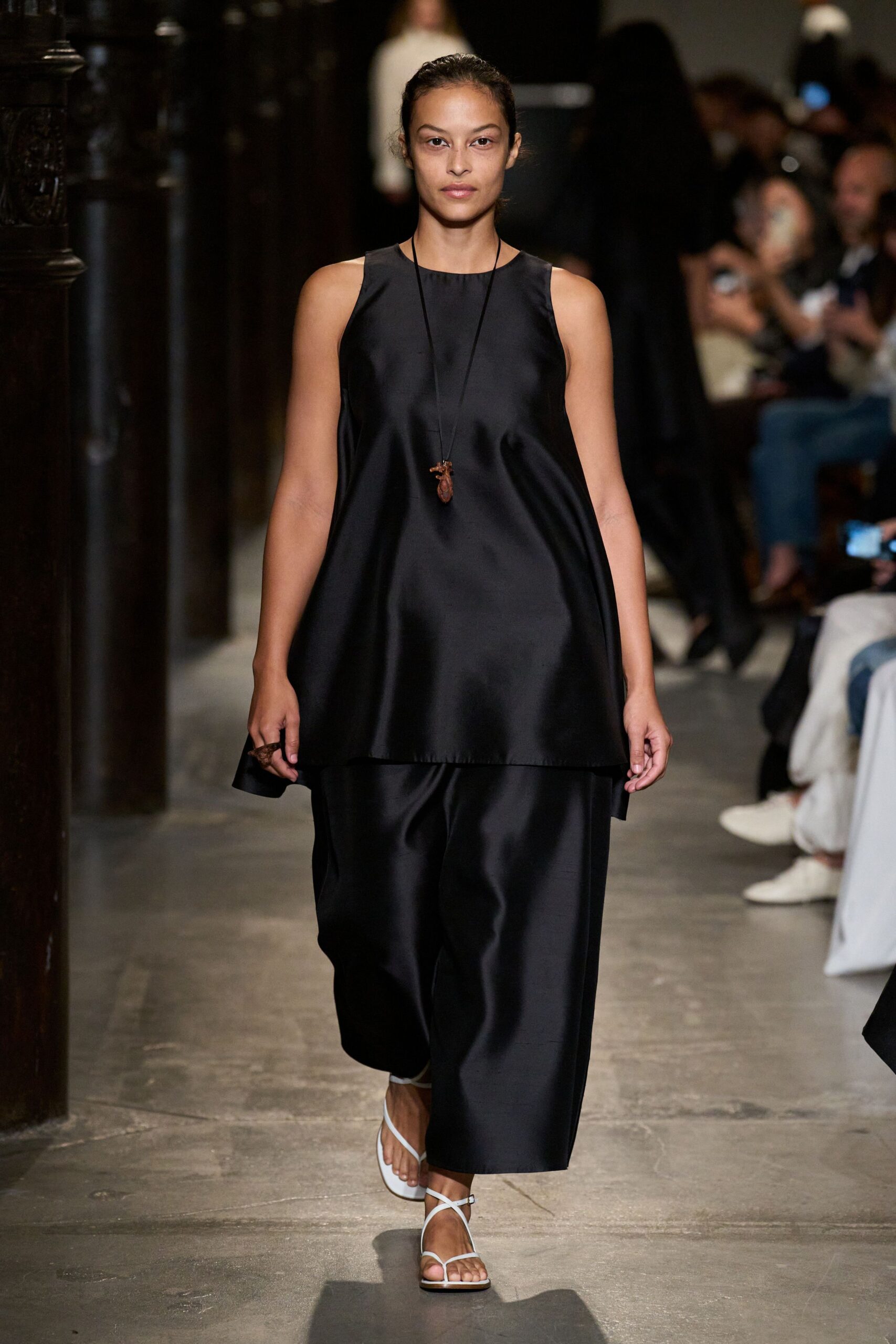 Black satin tunic cut wide from the armholes, hem dipping longer at back. Long cord necklace with a small carved brown pendant. Loose black satin trousers, cropped above the ankle. White thong sandals with thin crossed straps. Walking a quiet cloister corridor after the bells have gone.
