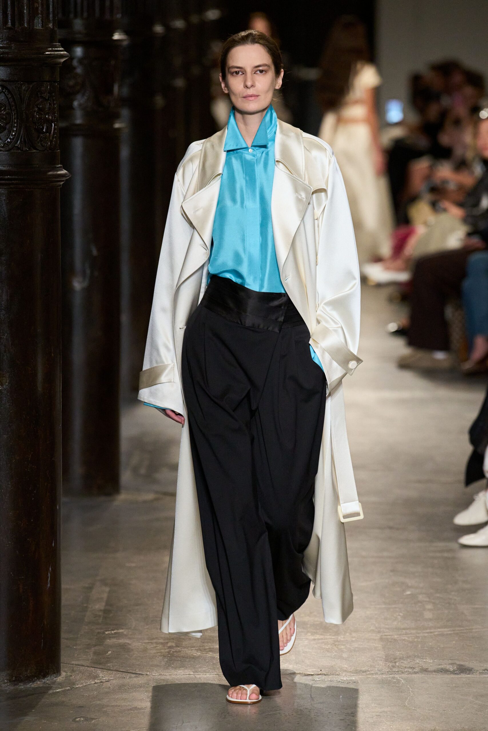 Ivory satin trench with oversized lapels and a dangling buckle tab. Turquoise satin blouse, pointed collar standing crisp above the coat. Black wide-leg trousers with a wrapped waistband and deep front pleats. White thong sandals. Walking through a hushed hotel corridor before the wedding guests wake.