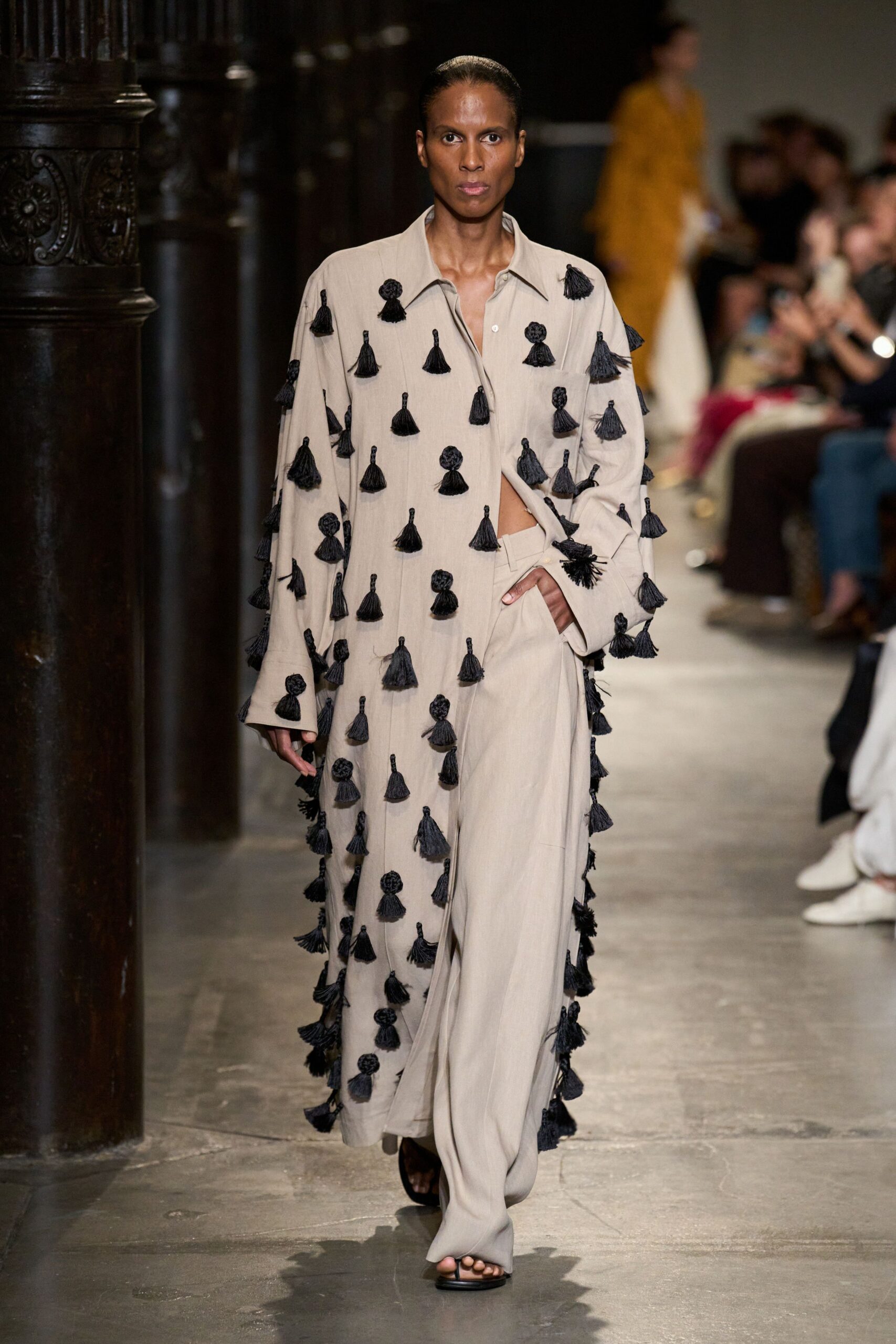 Oversized sand shirt scattered with black tassels, one placket corner kicked open. Loose matching trousers with broad pleats falling over the sandals. Black flip-flop sandals. Crossing a stone cloister after lunch, still carrying hotel room keys.
