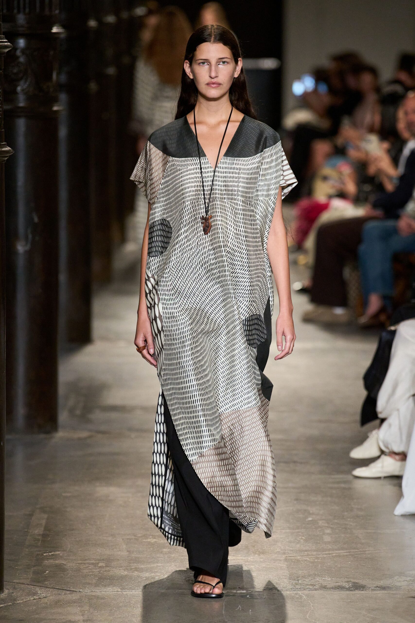 Black-and-white patchwork caftan, sheer grid panels interrupting dashed and dotted prints. Loose black trousers falling straight beneath the uneven hem. Long cord necklace with a small carved pendant. Chunky brown ring. Flat black thong sandals. Walking through a stone cloister after the heat has broken.