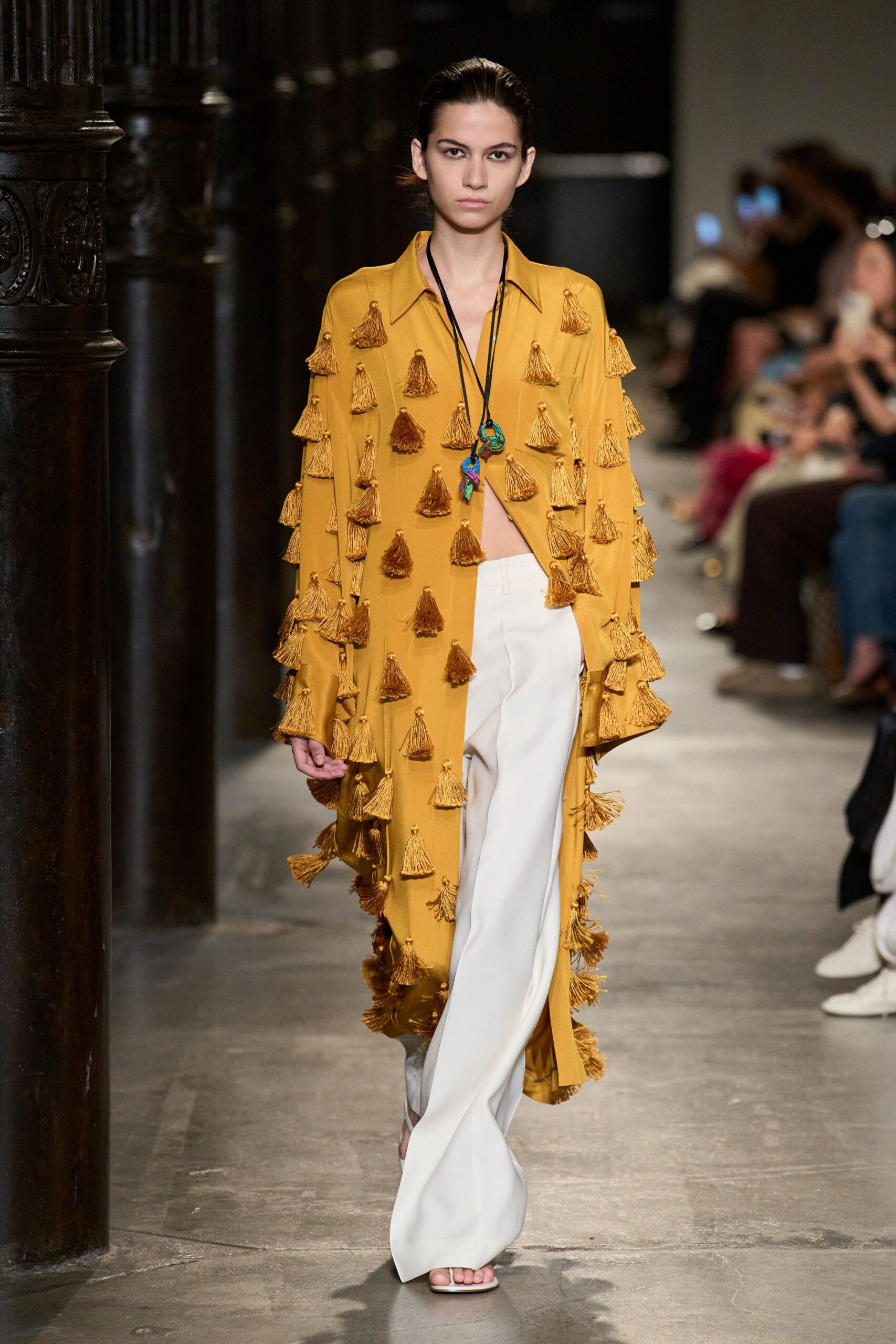 Mustard duster coat covered in swinging tassels, hem brushing open at center. Black cord necklace with bright beadlike charms hanging into the opening. Ivory flared trousers with a sharp crease and pooled break. Barely there white sandals. Crossing a tiled courtyard after lunch, hotel shutters still half closed.