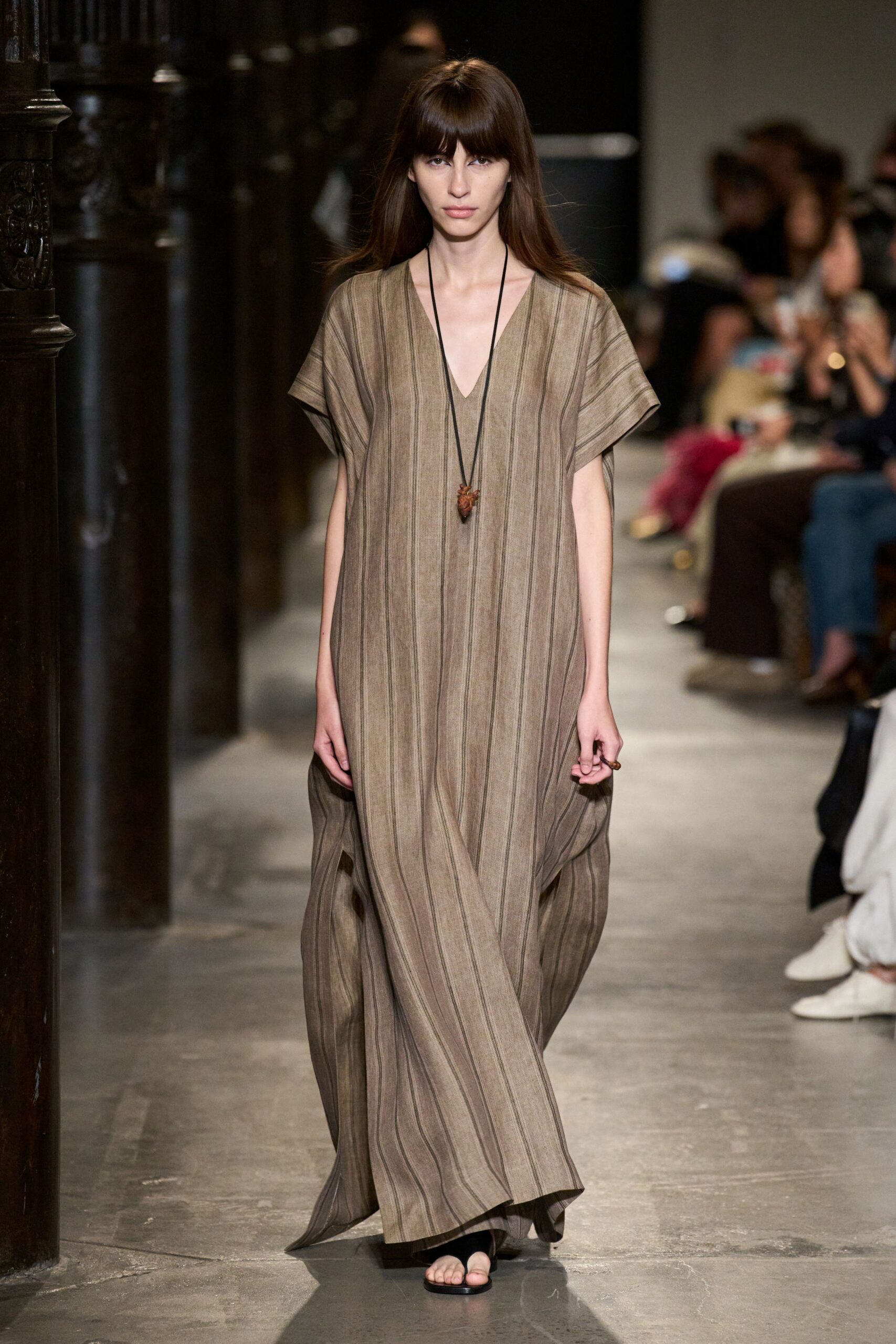 Taupe striped caftan dress in airy slub weave, deep V and cap sleeves. Long black cord with a carved seed pendant at the sternum. Flat black thong sandals, barely there under the sweeping hem. Crossing a cool stone cloister just after the noon bells.
