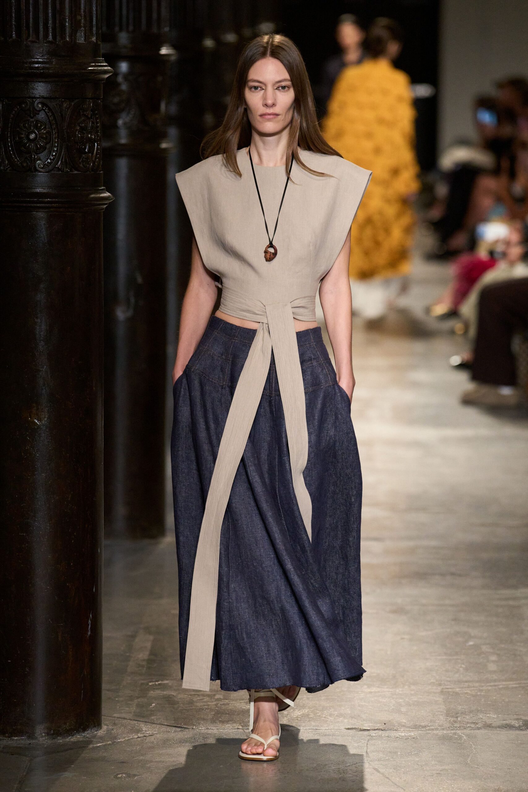 Oat linen top with winged shoulders and a long front tie. Small carved wooden pendant on a black cord. Dark indigo chambray skirt, deeply pleated with handsunk side pockets. White thong sandals with slim wraparound ties. Walking through a cloister courtyard before the flagstones give back the heat.