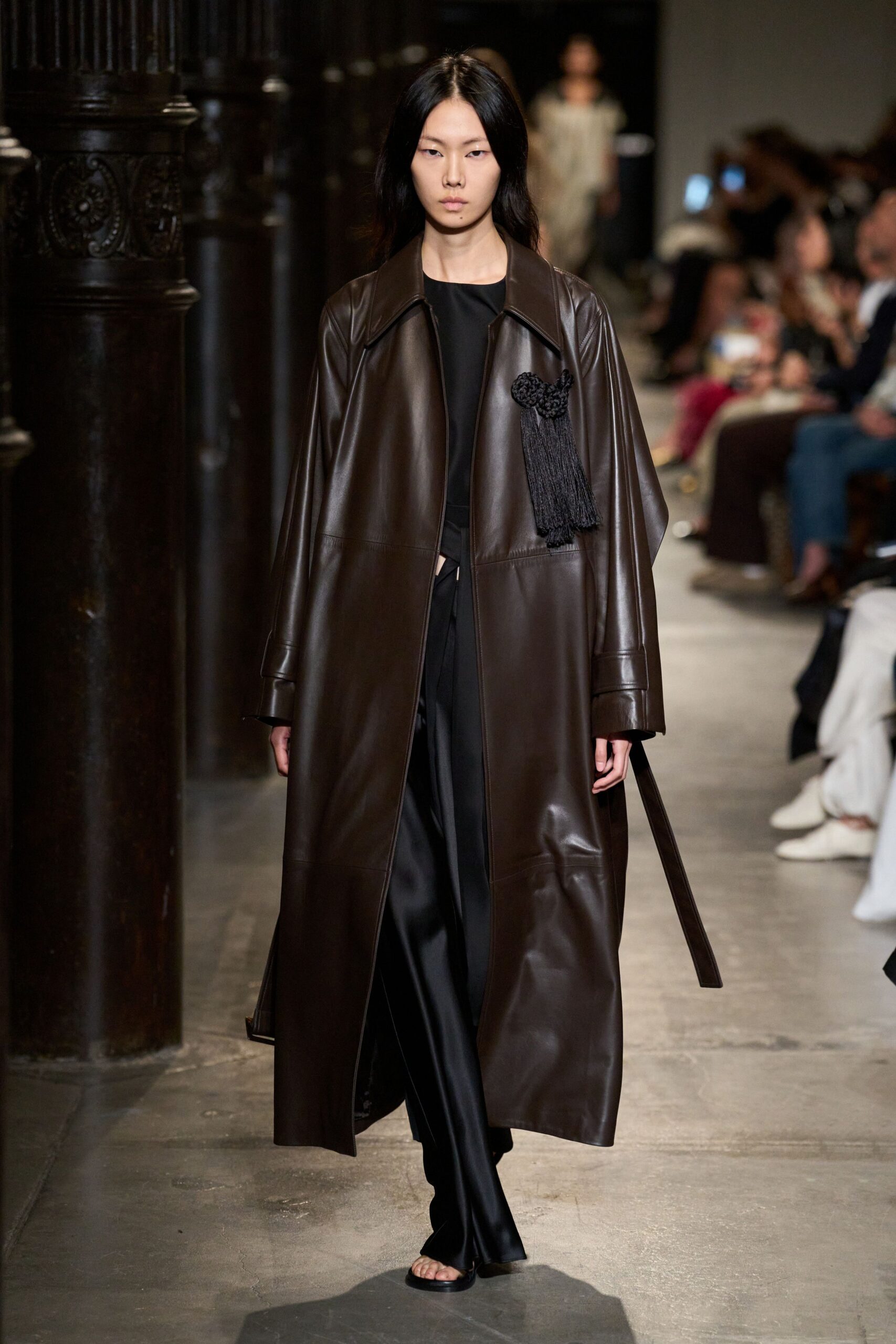 Long espresso leather coat with dropped shoulders and a black tassel cluster. Black crewneck top, plain and matte beneath the shine. Fluid black satin trousers, a small waist cutout peeking through. Flat black thong sandals. Walking through a stone cloister after rain, still carrying the evening with you.