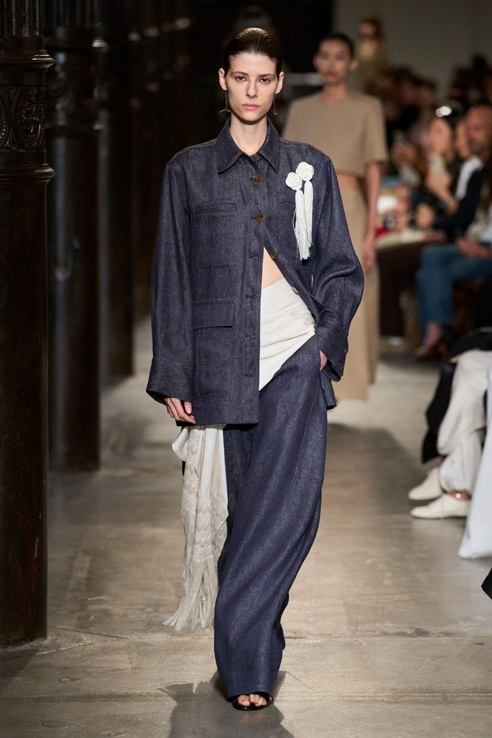Dark indigo denim jacket, brown buttons skewed open over one hip. Three white rosettes with a long tassel falling past the pocket. Ivory jersey wrapped low across the waist. Wide indigo denim trousers with a pooled break over black toes. Black open-toe slides. For crossing a stone cloister after a wedding has ended.