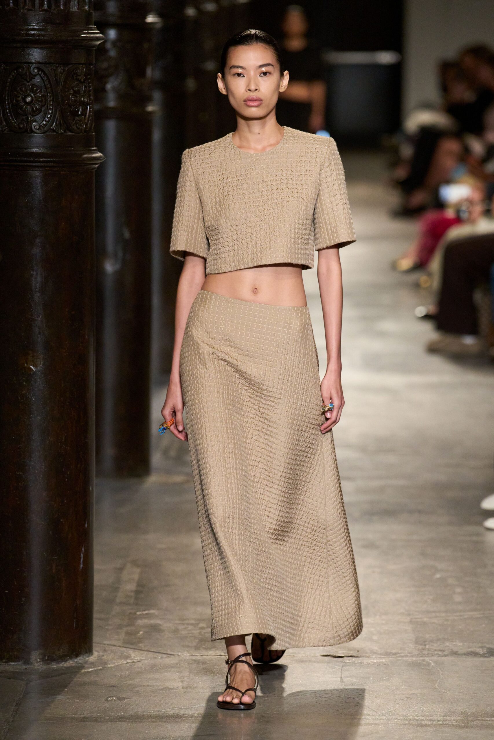 Sand-coloured basket-weave crop top with square shoulders and a clean hem. Matching bias-cut skirt, the gridded texture bending into a soft diagonal pull. Dark leather toe-loop sandals with thin crossing straps. Chunky resin rings in candy-bright colours. Crossing a cloister courtyard after lunch, stone still warm underfoot.