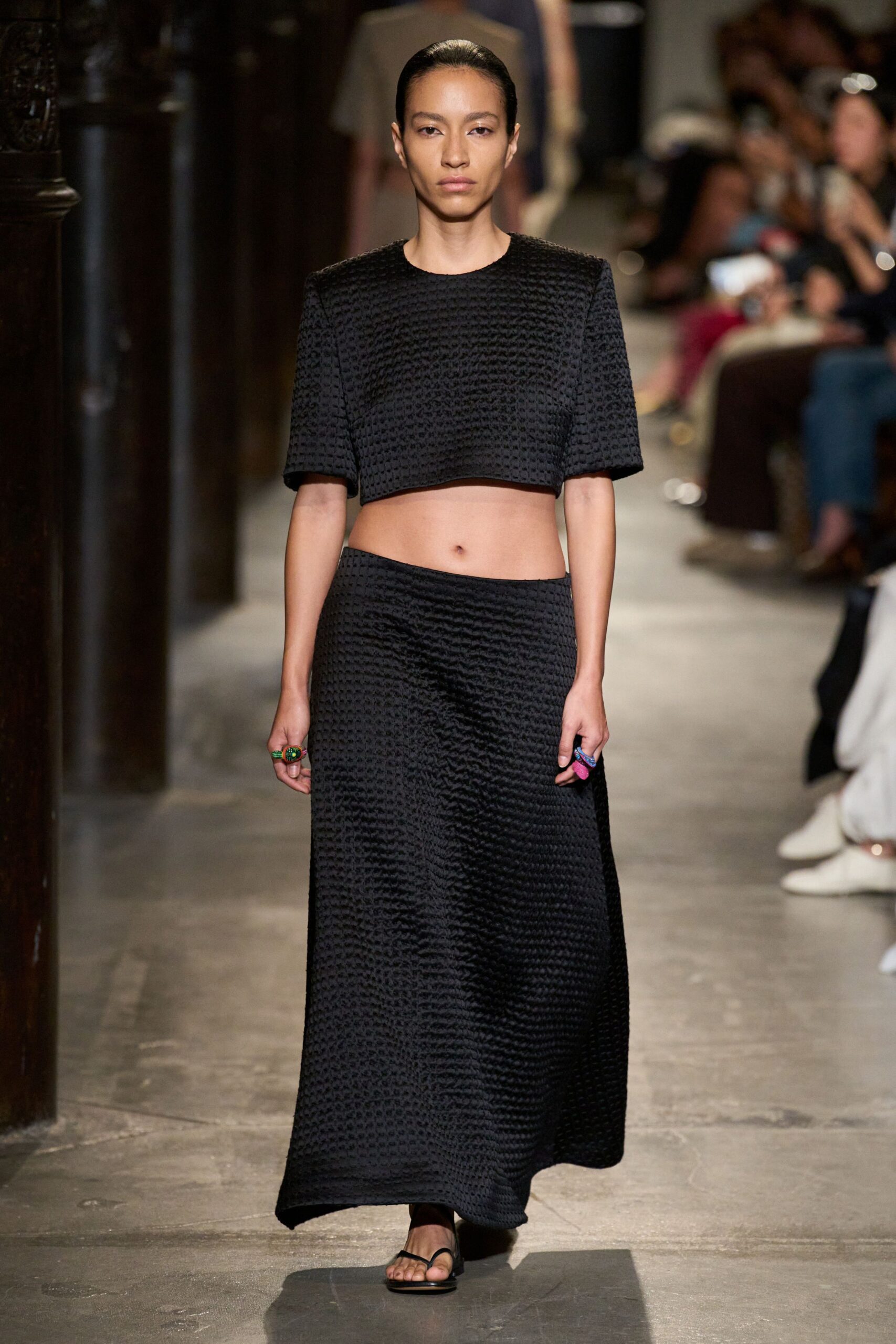 Black waffle-textured crop top with squared shoulders and a raw-looking hem. Matching ankle-length skirt in the same puckered weave, hanging slightly off-center. Flat black thong sandals. Chunky resin rings in grass green and bubblegum pink. Walking across cool church stone after the wedding guests have gone.