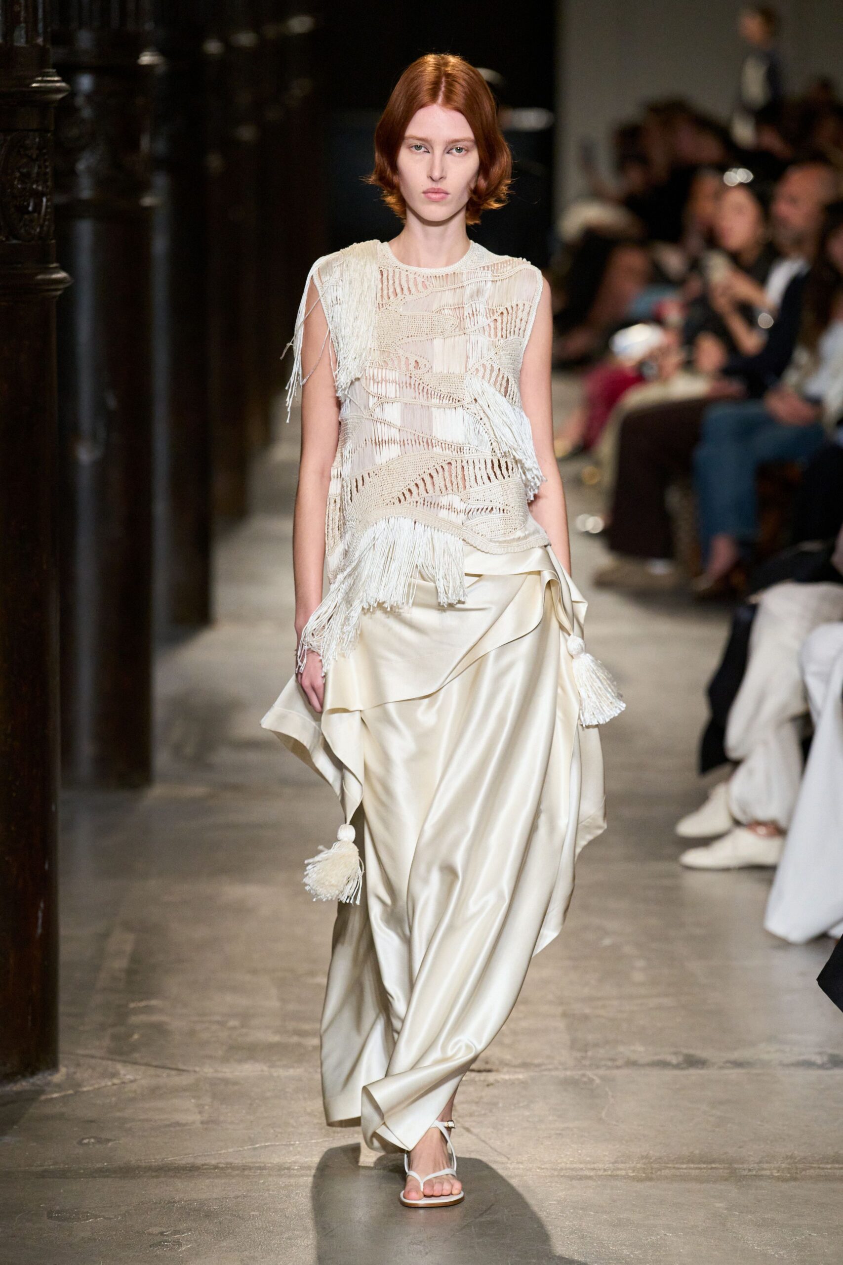 Ivory open-knit shell, laddered into wave bands with long fringe falling loose. Cream satin wrap skirt, swept into deep folds with tassels at each corner. White thong sandals with narrow straps. Crossing a cloister corridor after a wedding, shoes clicking on old stone.