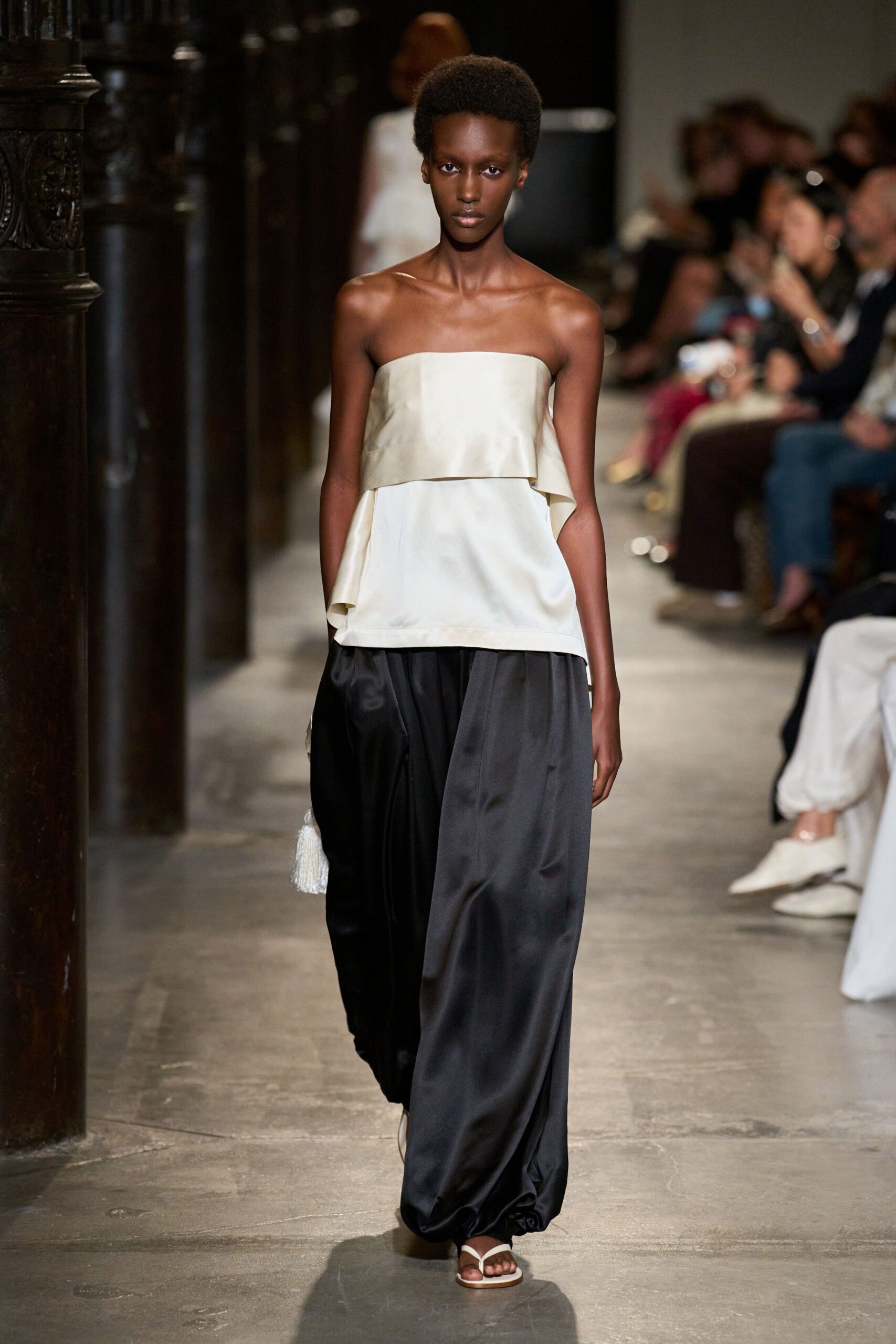 Ivory satin tube top with a loose overlay panel tipping at one side. Black satin wide-legs pooling into a soft gathered hem at the floor. White thong sandals with flat soles. Walking through a stone cloister after the wedding guests have gone.