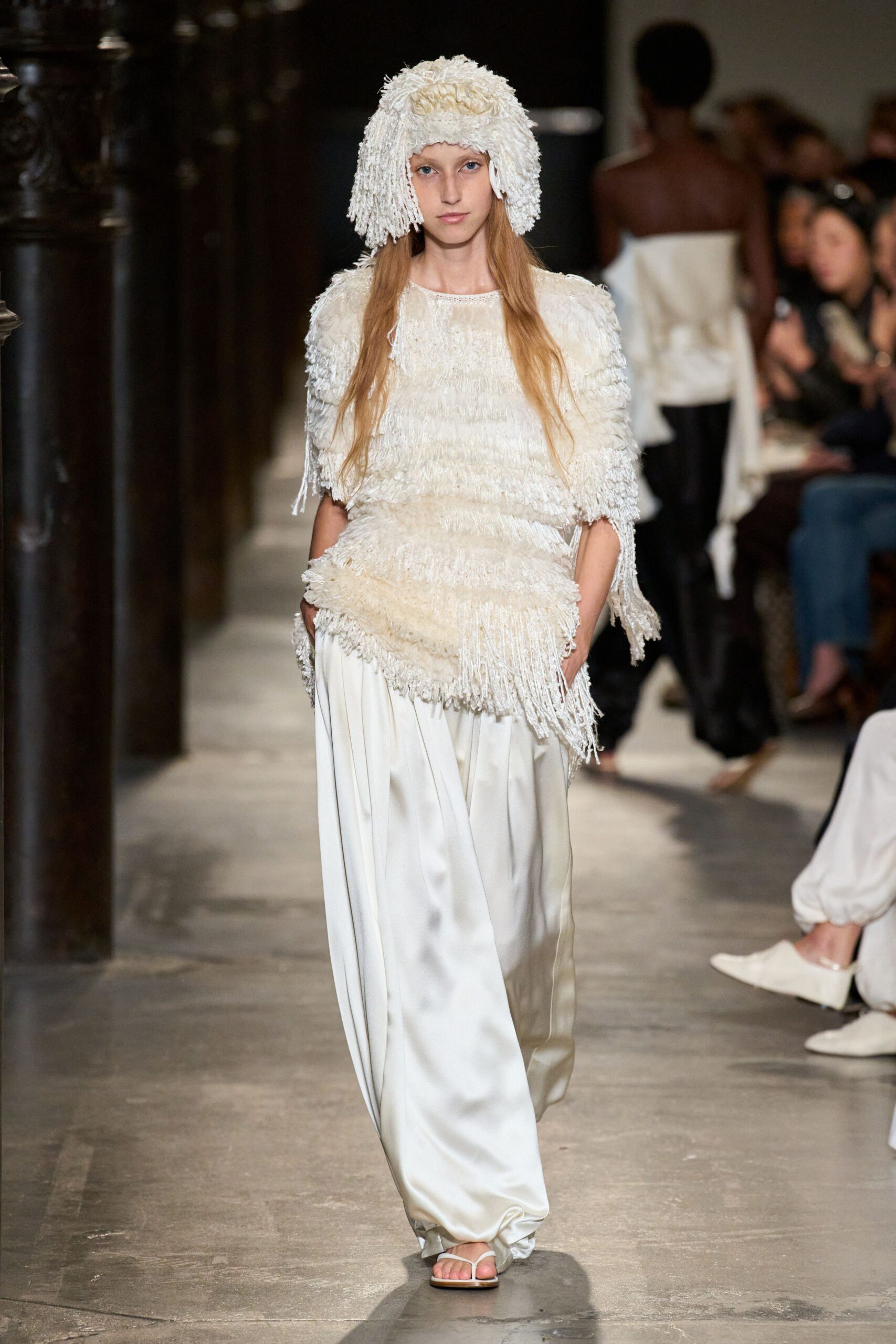 Cream tassel-knit cloche with a knotted crown and shaggy curtain brim. Ivory fringed top built in slanted tiers, one side hanging lower. Pearl satin wide-leg trousers pooling into soft creases over the foot. White thong sandals. Crossing a monastery courtyard just after the stone dries from rain.