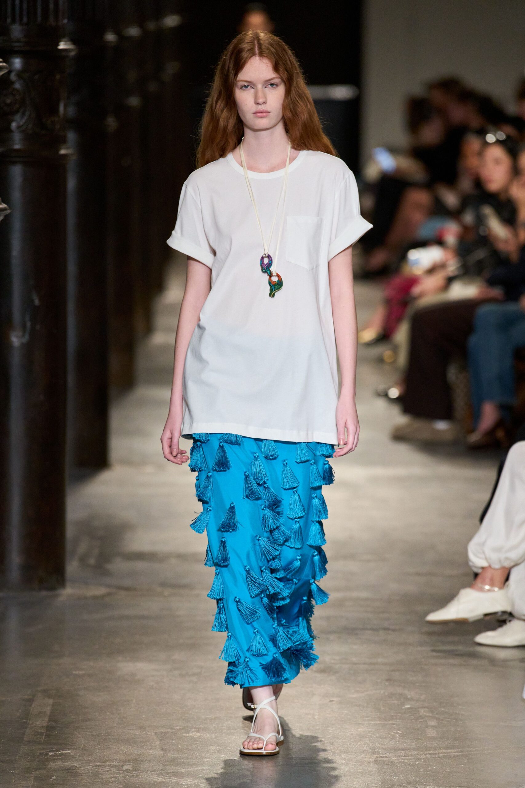 Oversized white jersey tee with rolled cuffs and a chest pocket. Long cord necklace hung with two enamelled paisley pendants. Turquoise column skirt densely covered in swinging thread tassels. White thong sandals with a slim ankle strap. Walking back from the beach bar with salt still drying on your shoulders.
