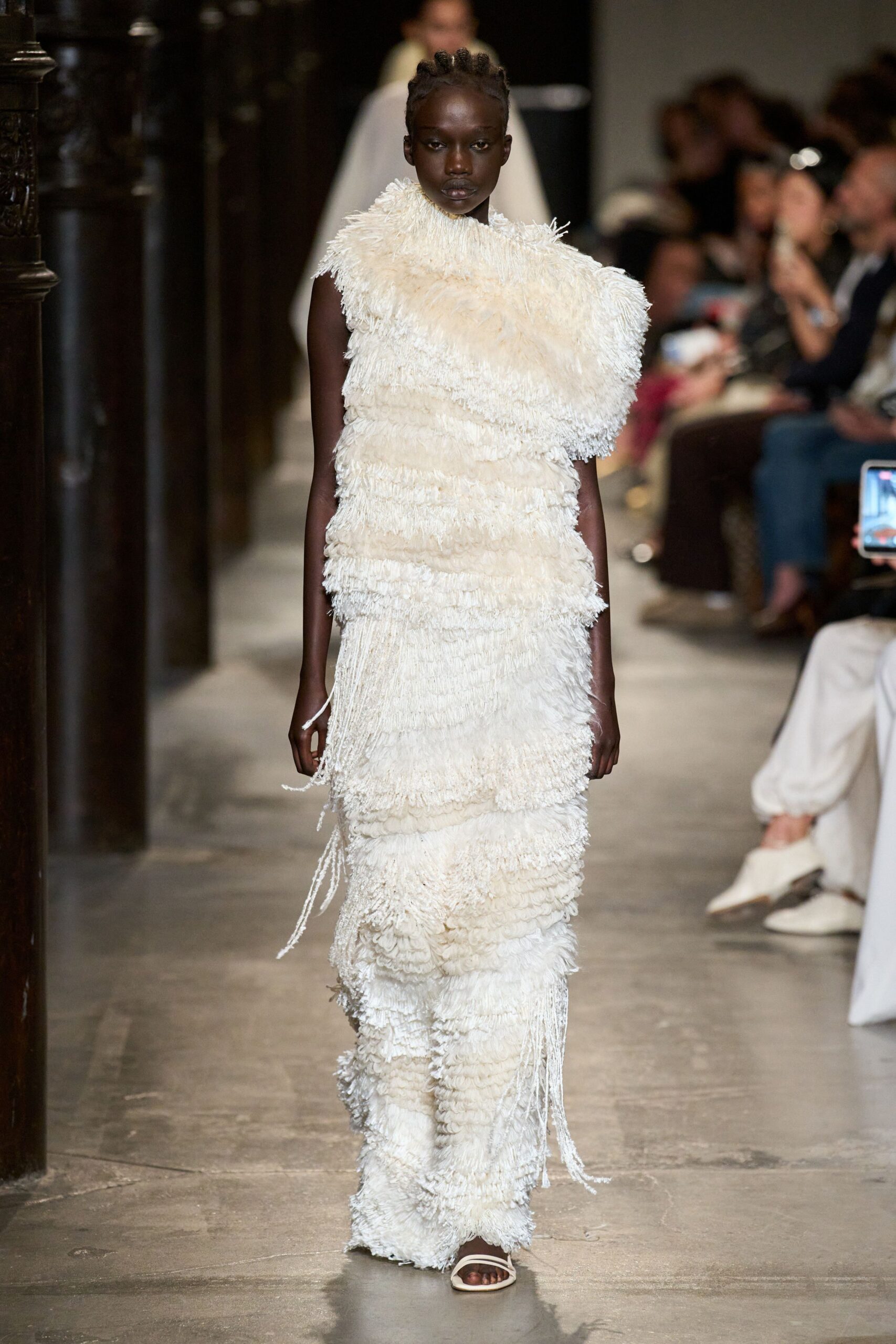Ivory column dress built in dense shaggy fringe, one shoulder swelling into a high cowl. Barely-there ivory slide sandals. Crossing a stone cloister just after the bells have finished.