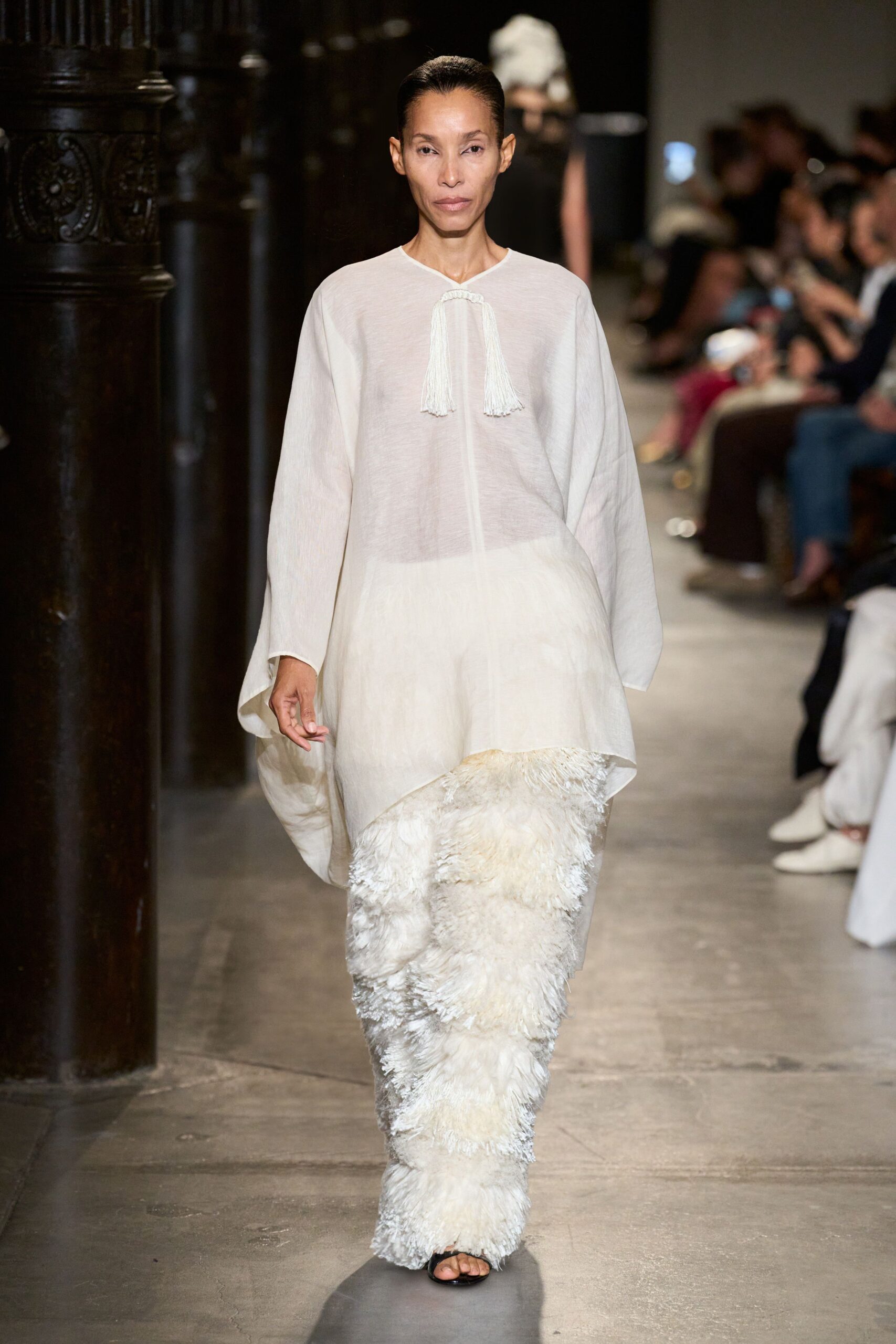 Sheer ivory tunic with dropped shoulders and two long tassels at the slit neck. Ivory trousers buried under shaggy featherlike fringe, brushing over the foot. Black open-toe sandals just peeking out. Crossing a hushed cloister courtyard after the bells have finished.