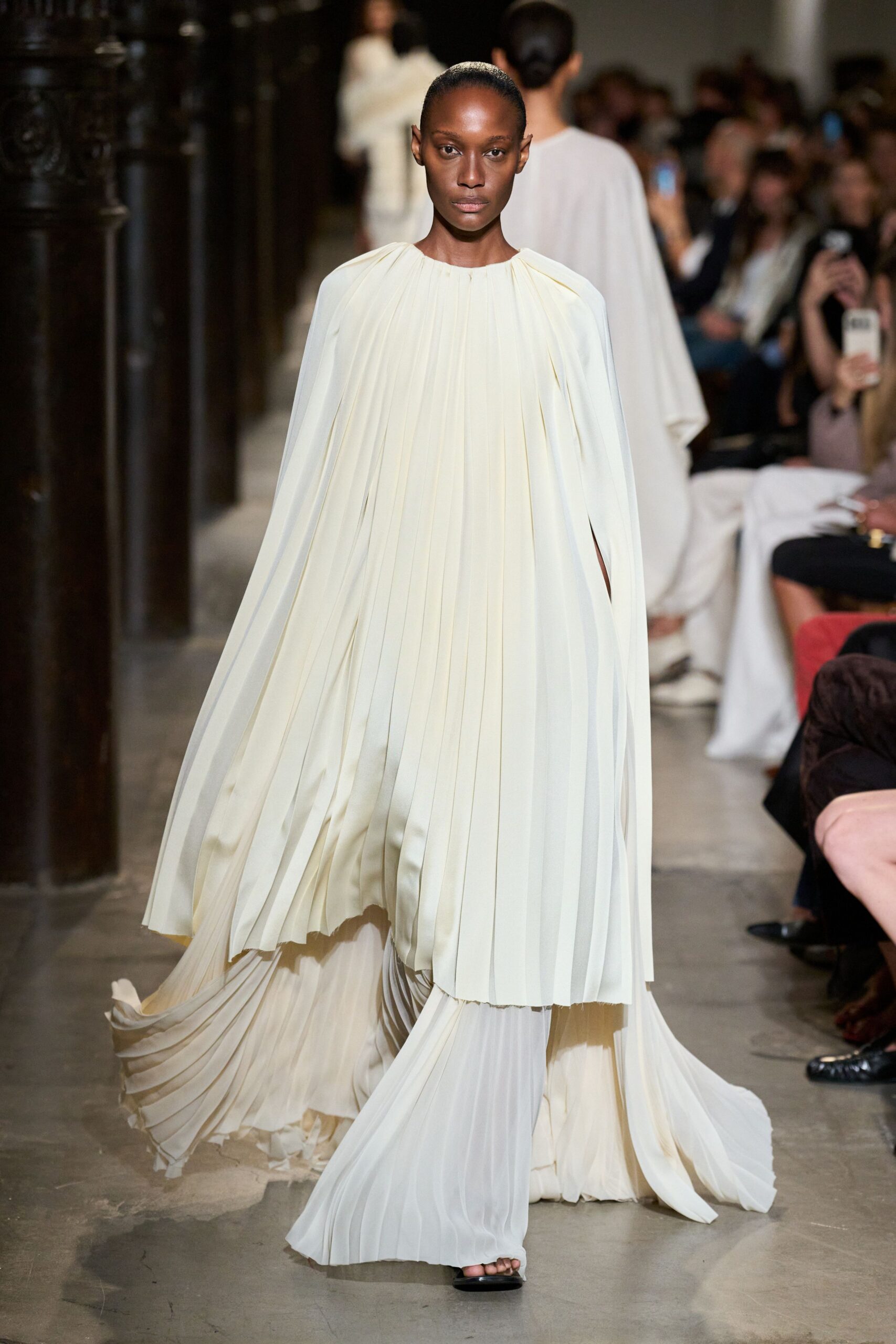 Ivory chiffon gown cut in knife pleats, gathered tight at the neck. Cape sleeves slit open at the arms, falling in long panels. Matching pleated trousers pooling wide over a black open sandal. Crossing a marble cloister just after the bells stopped.