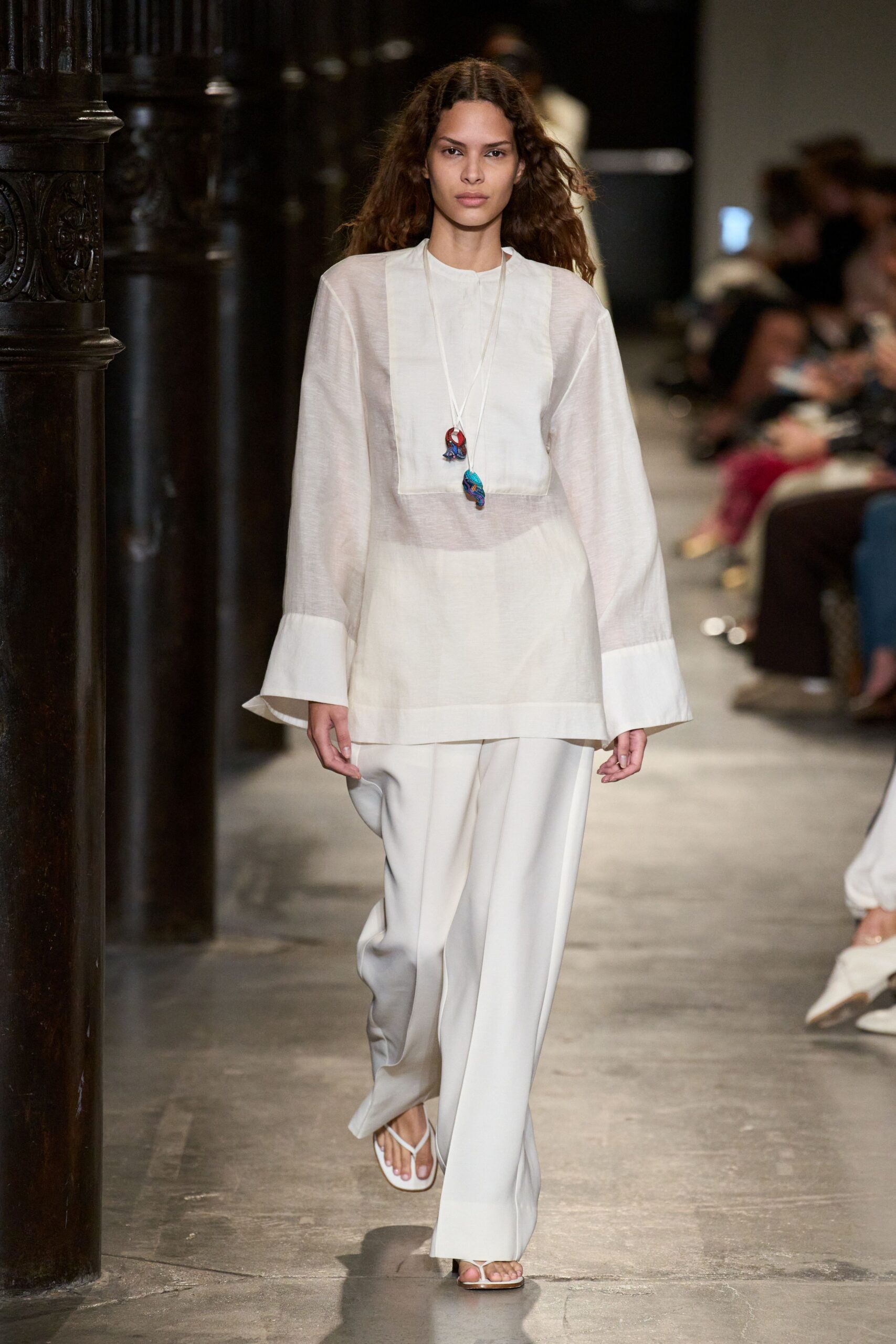 Sheer ivory tunic with a square bib panel and flared cuffs. Long cord necklace with two small glass pendants hanging off center. Wide ivory trousers in fluid crepe, breaking softly over the foot. White thong sandals. Walking through a cloister arcade after the bells, heat still in the stone.