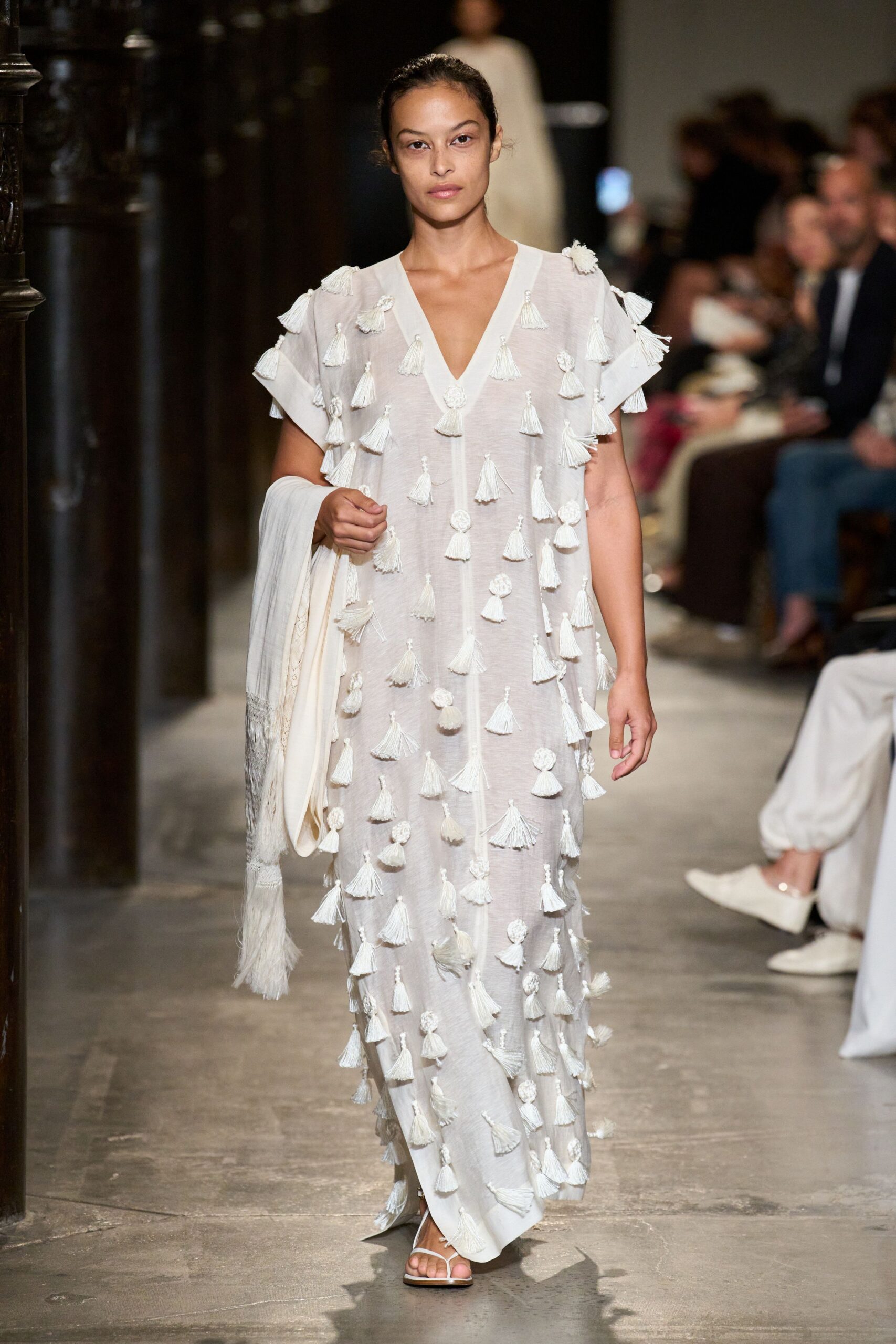 Sheer ivory caftan with a deep V and tassels knotted all over. Soft ivory wrap ending in long fringe and narrow woven borders. Flat white thong sandals. Crossing a stone cloister just after the heat breaks.