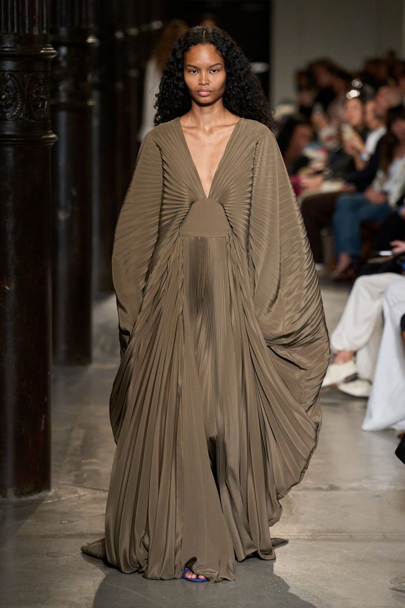 Taupe jersey gown with knife-pleated wings collapsing into pooled folds. Cobalt thong sandals peeking from the dragging hem. Crossing a stone cloister just before the bells start.