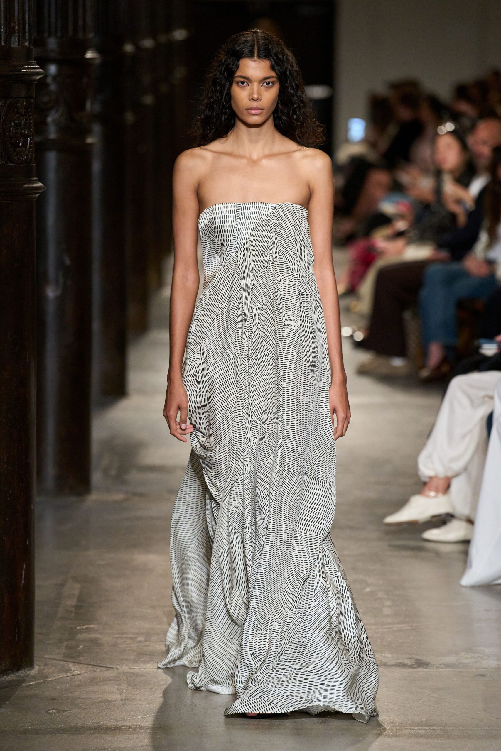 Strapless ivory gown in airy crinkled cloth, black dash print swirling over deep draped folds. Crossing a tiled courtyard just after the hotel wedding has emptied out.