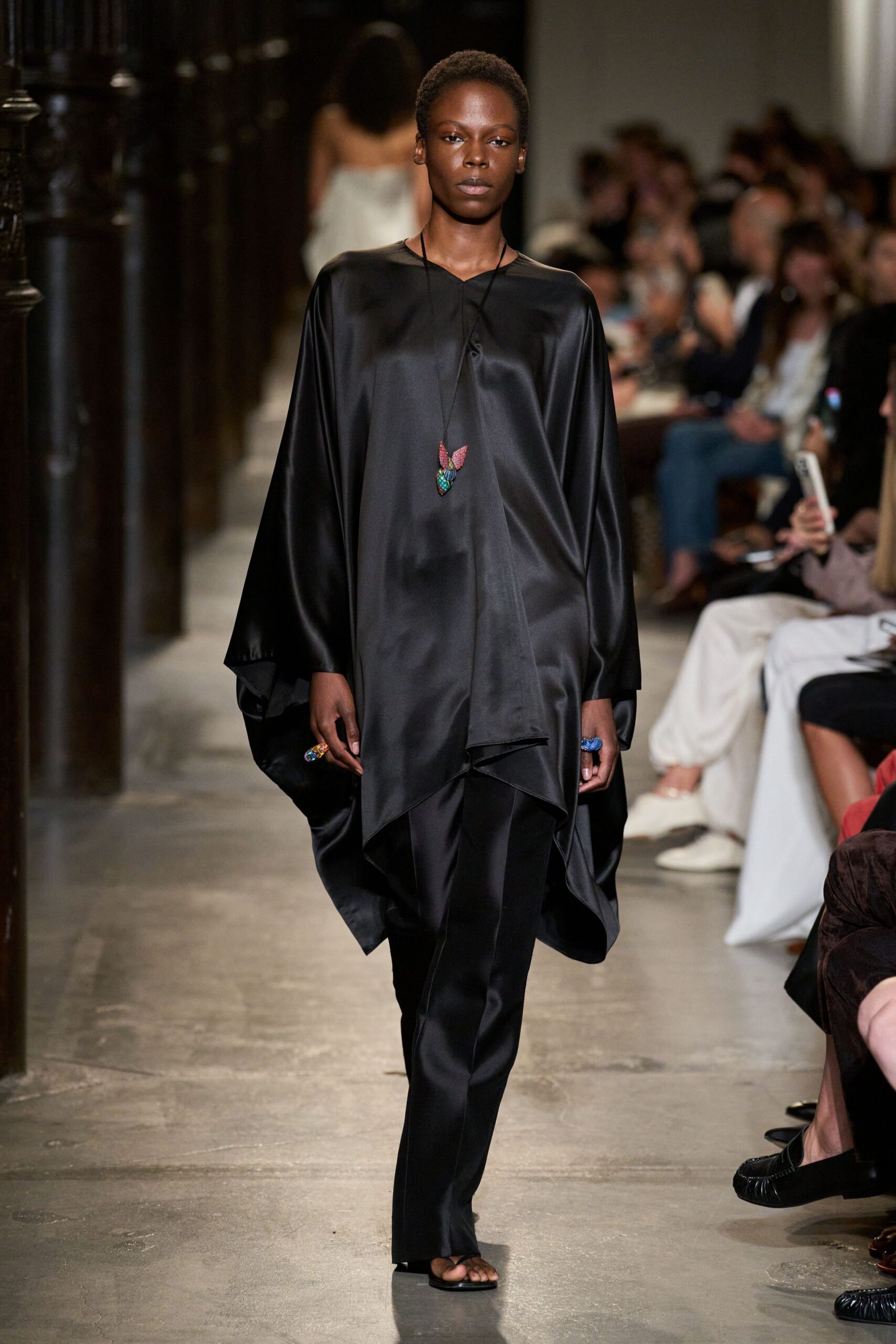 Black satin tunic cut in a loose caftan shape, hem falling to points. Long cord necklace with a small beaded leaf pendant. Chunky blue and multicolored rings on both hands. Slim black satin trousers breaking lightly over the foot. Flat black thong sandals. Crossing a museum atrium after hours, floor still warm from the sun.