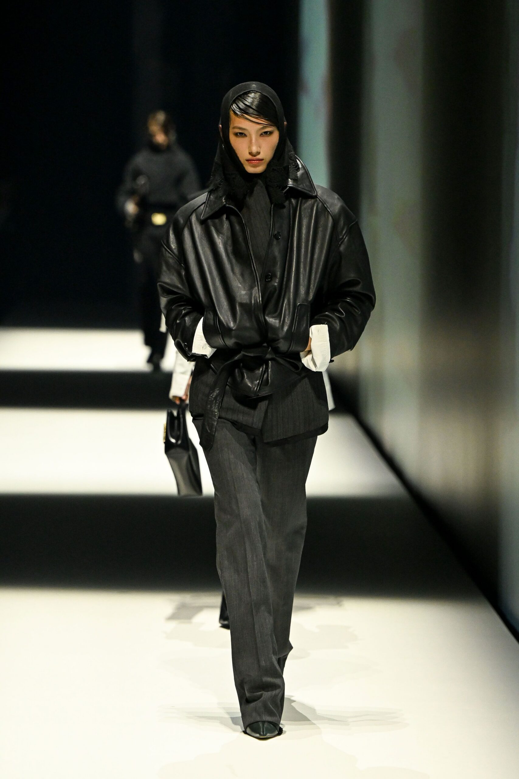 Black jersey headscarf framing slick side-swept bangs. Oversized black leather jacket, tied low with a long self belt. White cuffs jutting past the sleeves. Charcoal pinstripe trousers falling straight over the shoe. Square-toe black boots peeking from the hem. Walking into a courthouse lobby before the radiators have warmed up.