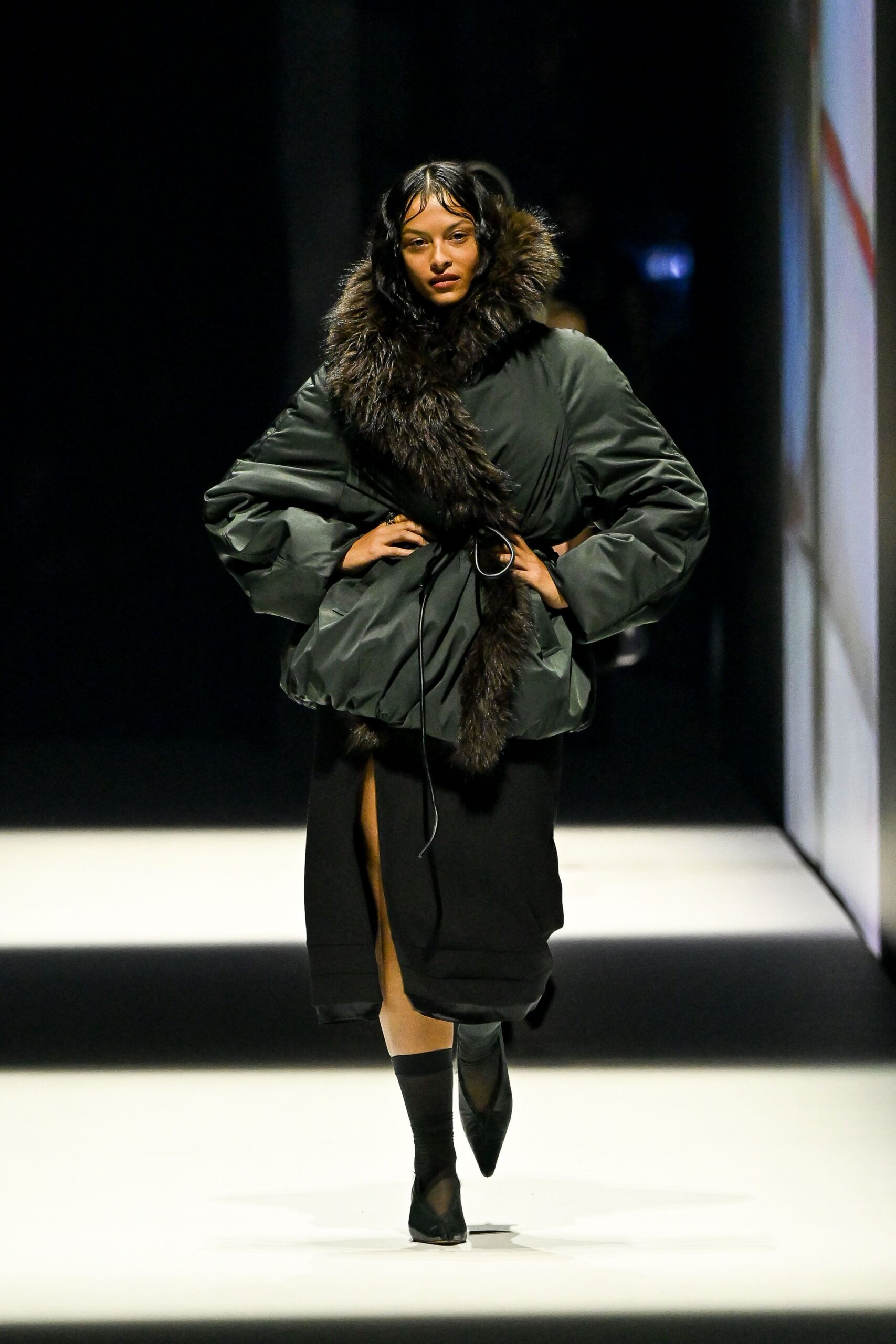 Charcoal puffed coat cinched with a thin cord through a metal ring. Dark shaggy fur wrapped high at the throat, one tail hanging long. Black skirt split high at the front with a satin-faced hem. Sheer black sock boots with pointed toes. Whatever you wear crossing the hotel forecourt after midnight in hard wind.