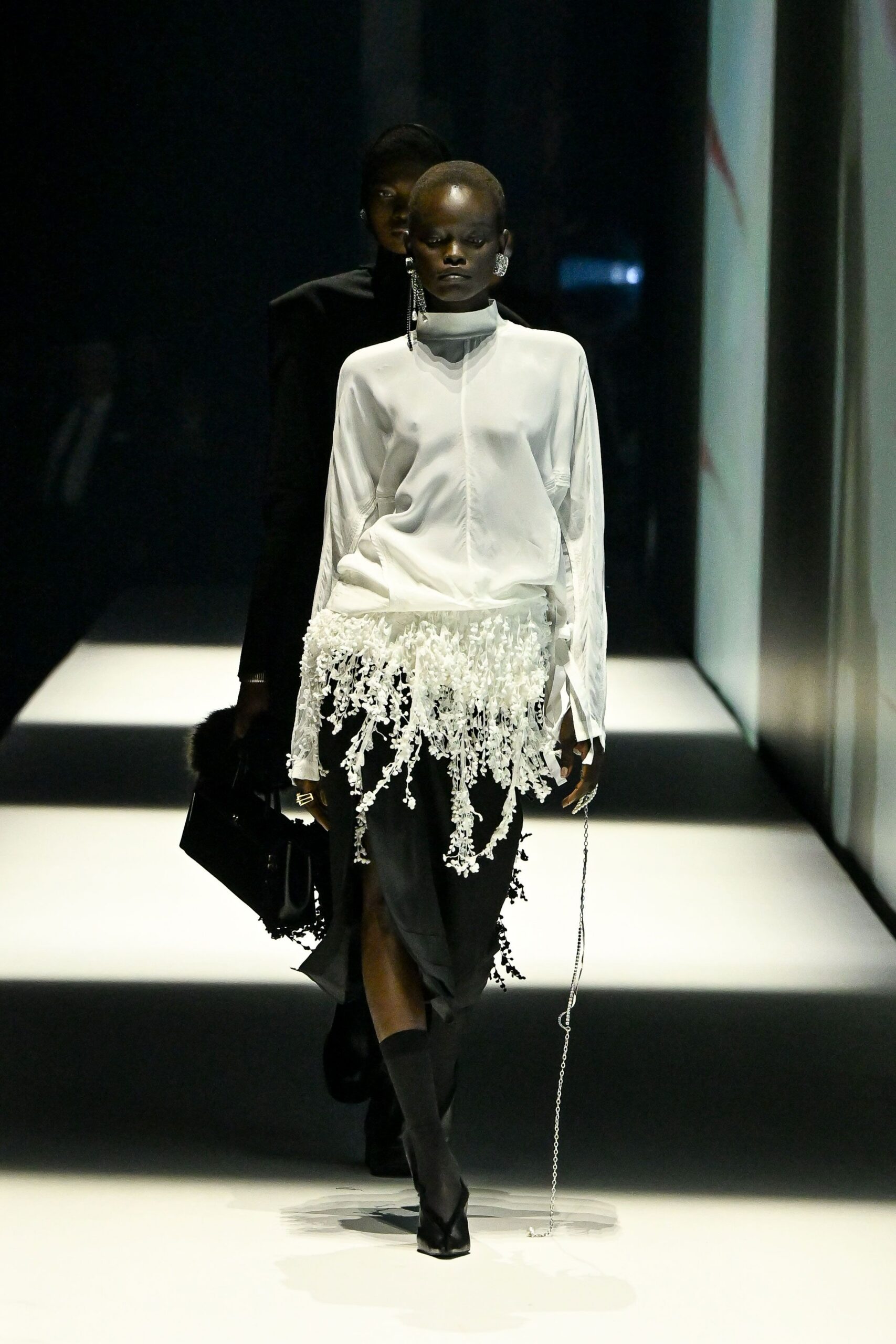 Ivory satin blouse with a soft funnel neck and gathered front seam. Black draped skirt, wrapped in dangling white floral fringe. Mismatched crystal drop earrings. Angular silver ring. Black top-handle bag with furry trim. Black sock boots with split toes. Walking through a hotel corridor after the wedding dinner emptied out.