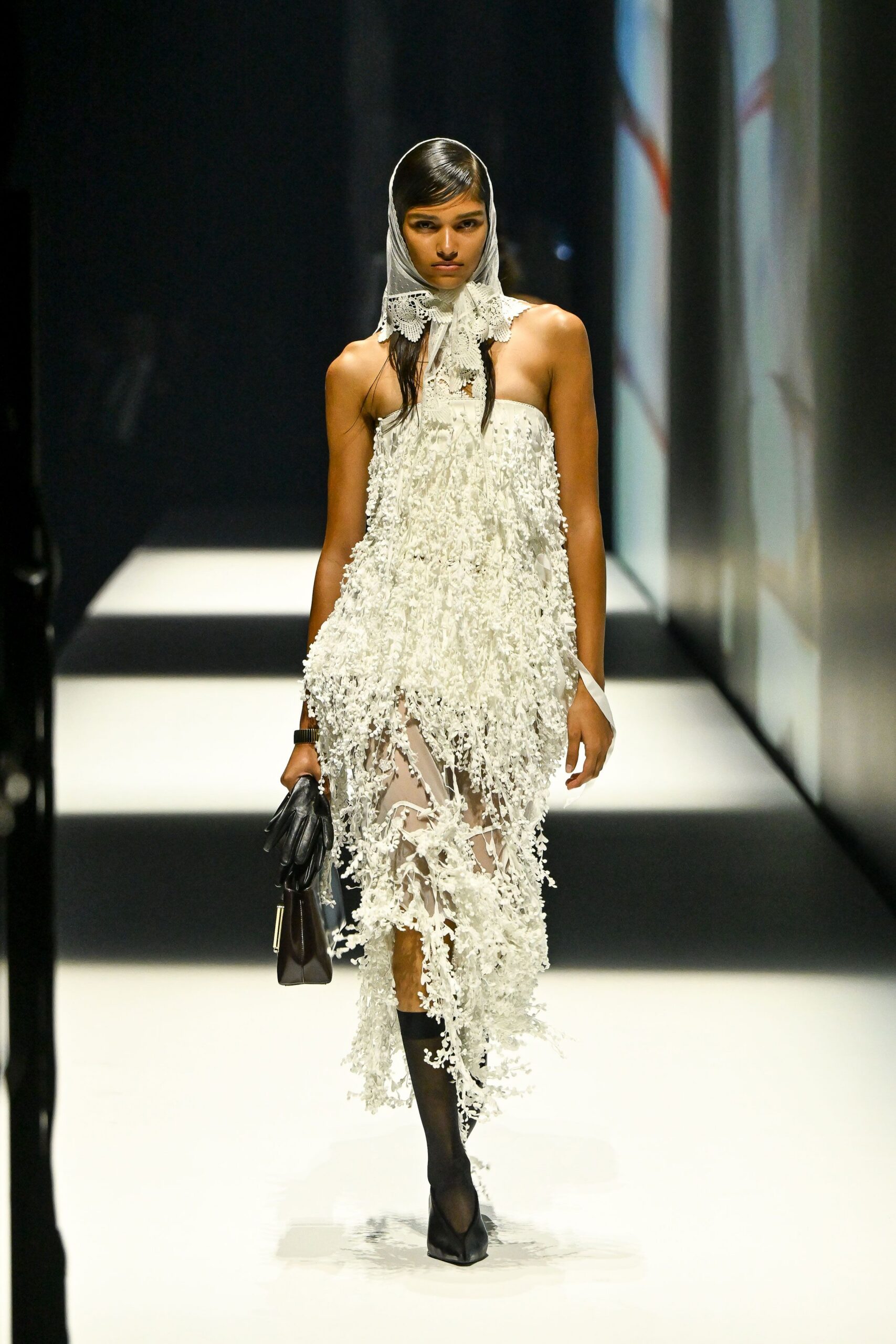 White scarf with a scalloped lace bib tied under the chin. Strapless ivory dress drowned in dangling floral appliqué over sheer mesh. Single black leather glove. Boxy dark satchel with a long metal clasp. Black knee-high sheer stockings. Square-toe black pumps. Church basement wedding, cake crumbs already ground into the floor.