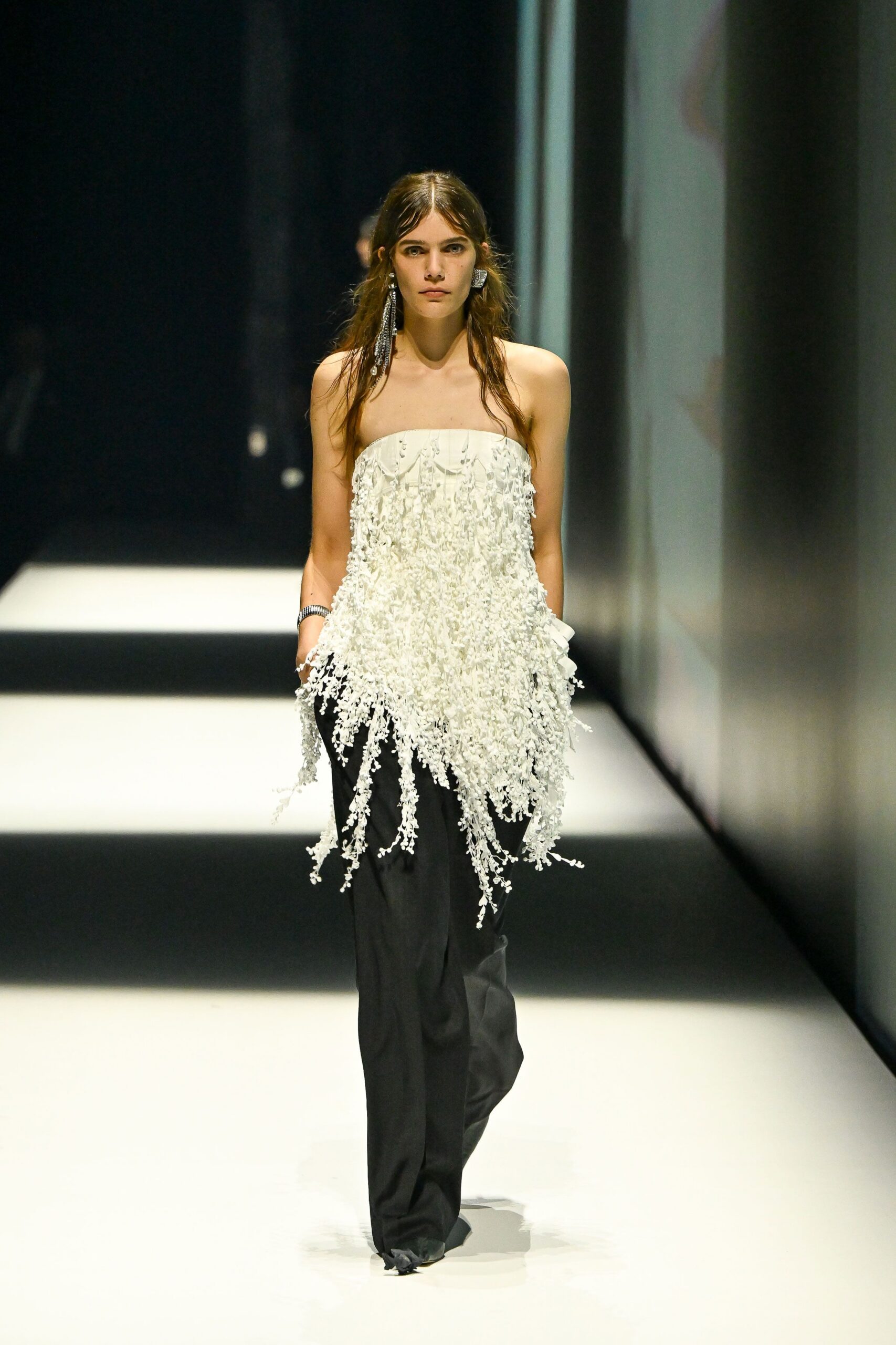 Mismatched silver earrings, one a long rhinestone fringe. Strapless ivory top densely crusted with dangling floral paillettes. Black wide-leg trousers breaking heavily over the shoe. Coiled silver cuff at one wrist. Crossing a hotel lobby after midnight with confetti still caught on you.