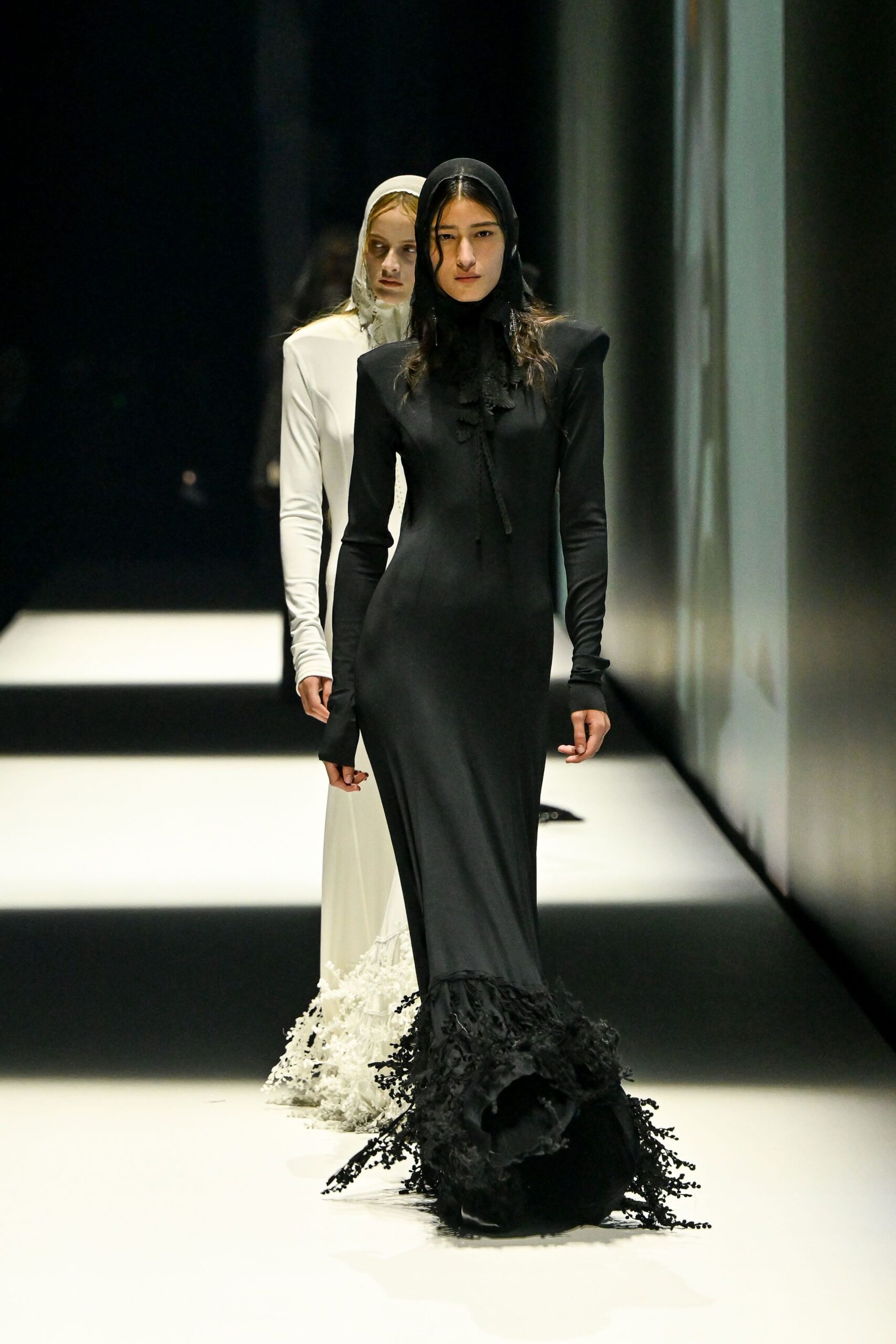 Black stretch hood framing the face close, one wet curl left loose. Long black jersey gown with sharp padded shoulders and thumbhole sleeves. Narrow ruffled tie falling off-center from the throat. Dense black fringe erupts at the train like seaweed. Crossing a hotel atrium before dawn, floor polish still tacky underfoot.