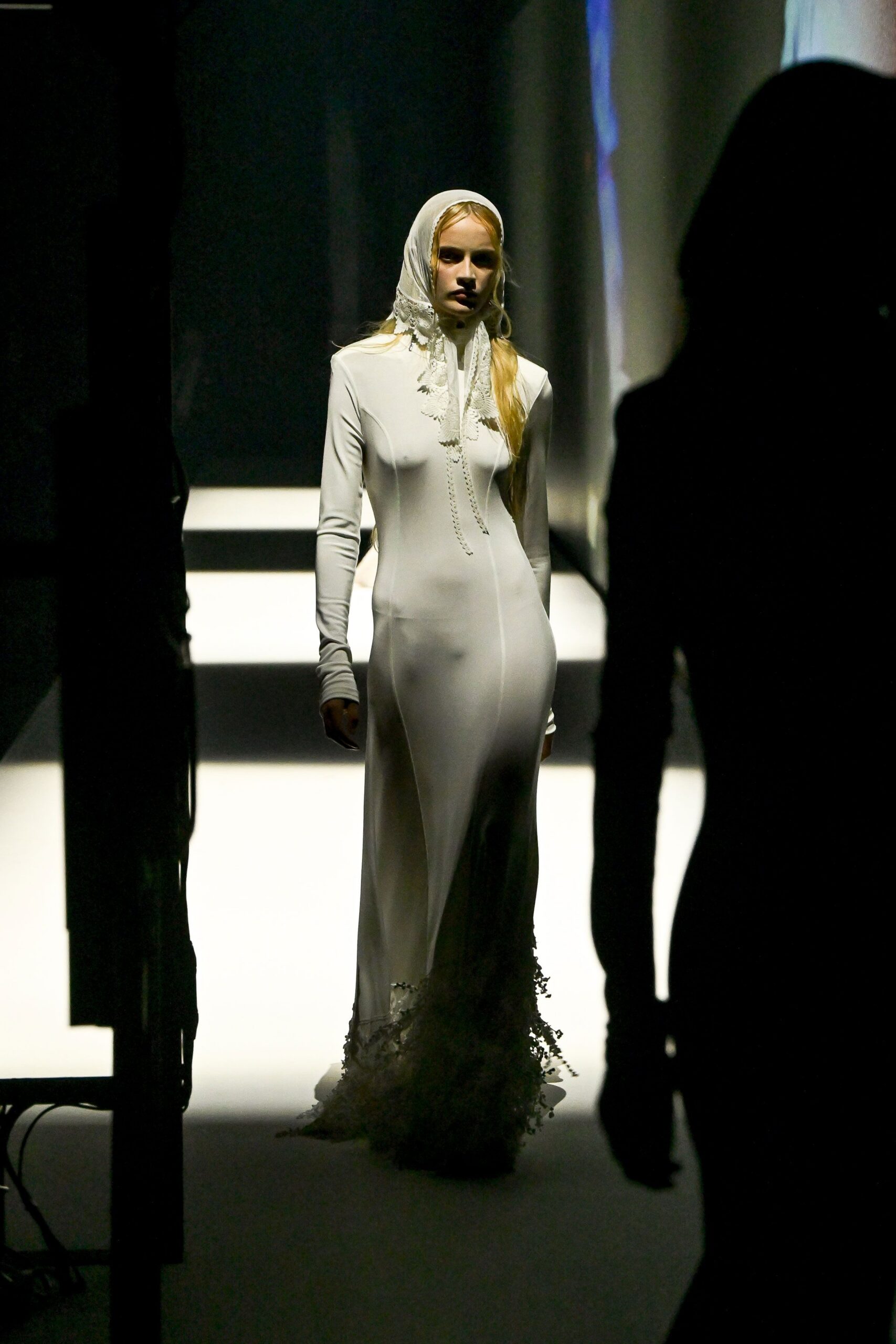 Ivory scarf edged in scalloped lace, tied close under the chin. White stretch column gown with a molded turtleneck and lace-frayed train. Crossing the chapel hall after everyone else has already gone outside.