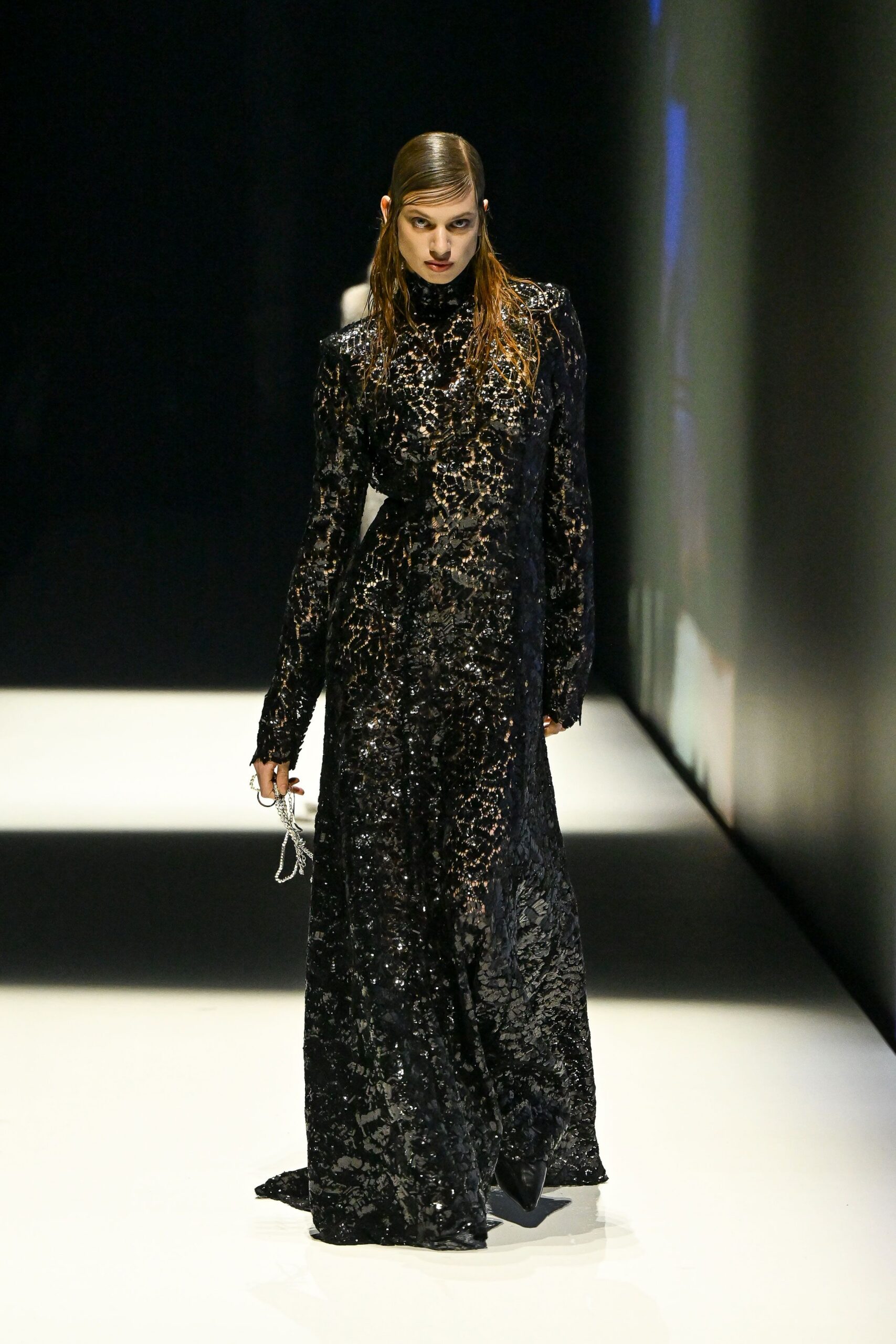 Black high-neck gown in glossy floral lace, sleeves ending past the knuckles. Pointed black boots disappearing under the pooling hem. Silver mesh pouch dangling from two finger rings. Whatever you wear home when the hotel bar carpet is still wet from cleaning.