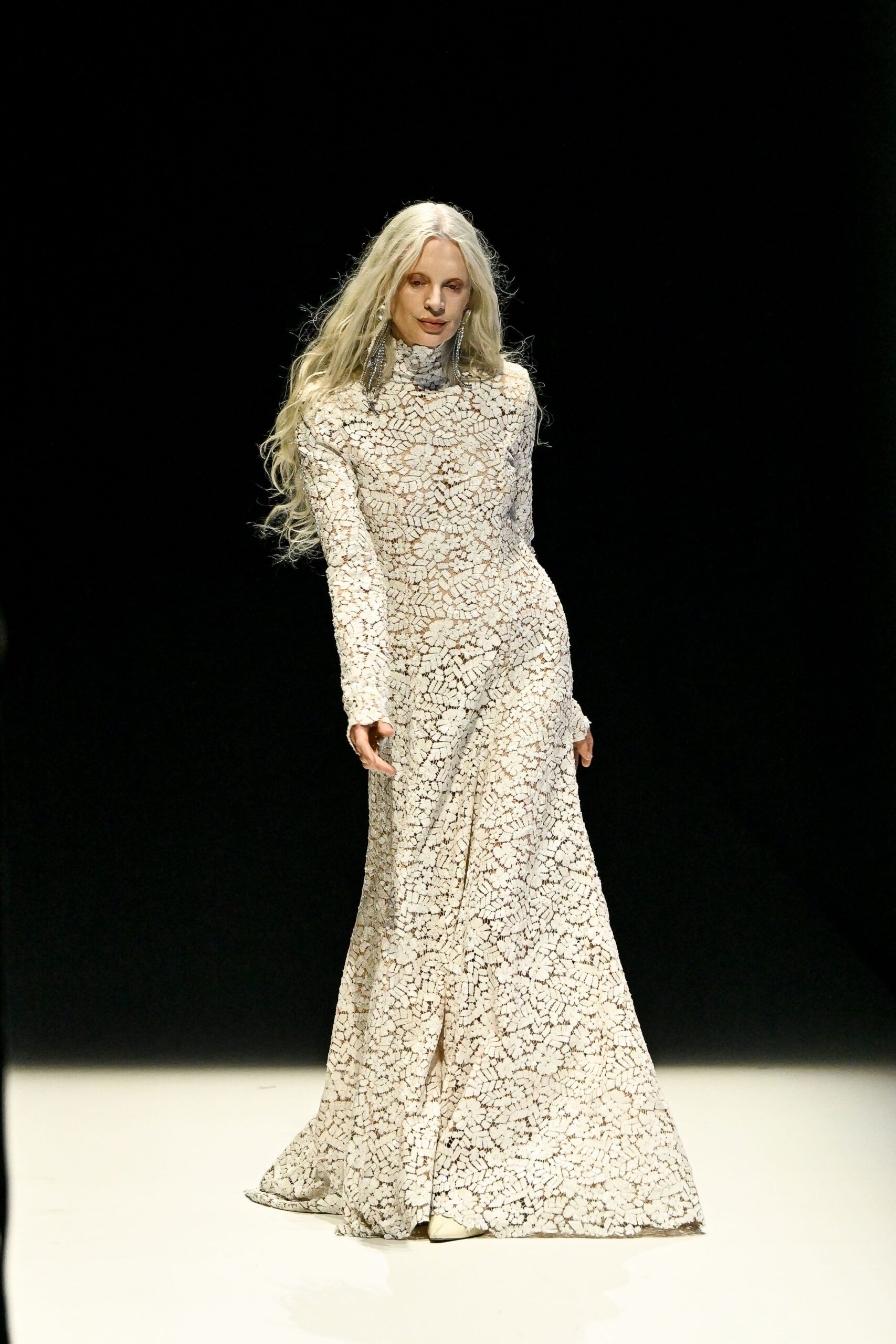 Long crystal fringe earrings brushing the jaw on both sides. Ivory high-neck lace gown, fitted through hip then loosening to a pooled hem. Pointed ivory boots barely peeking from the front slit. Walking across a museum atrium after hours, marble floor still holding the day's cold.