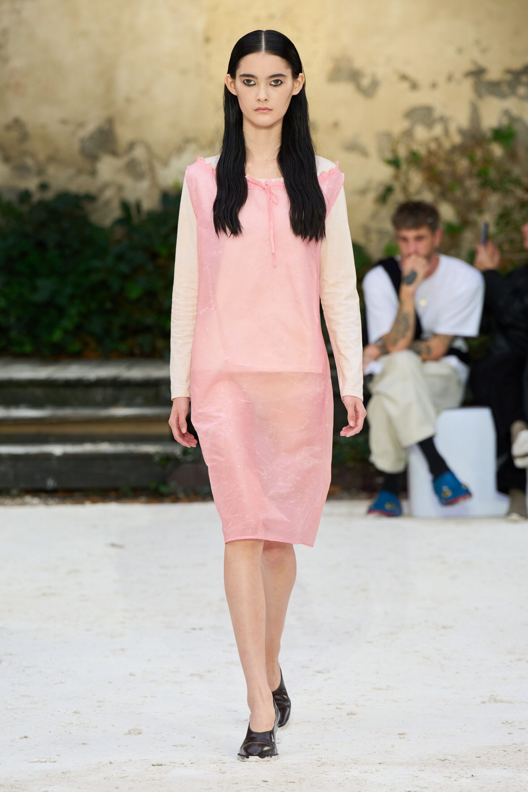 Pale pink translucent shell with a frayed neckline and dangling tie. Cream jersey sleeves showing underneath, close and almost sheer. Matching pink pencil skirt in cloudy crinkled plastic-like voile. Black leather flats with squared toes. Crossing a sunblown courtyard after the fountain has been turned off.